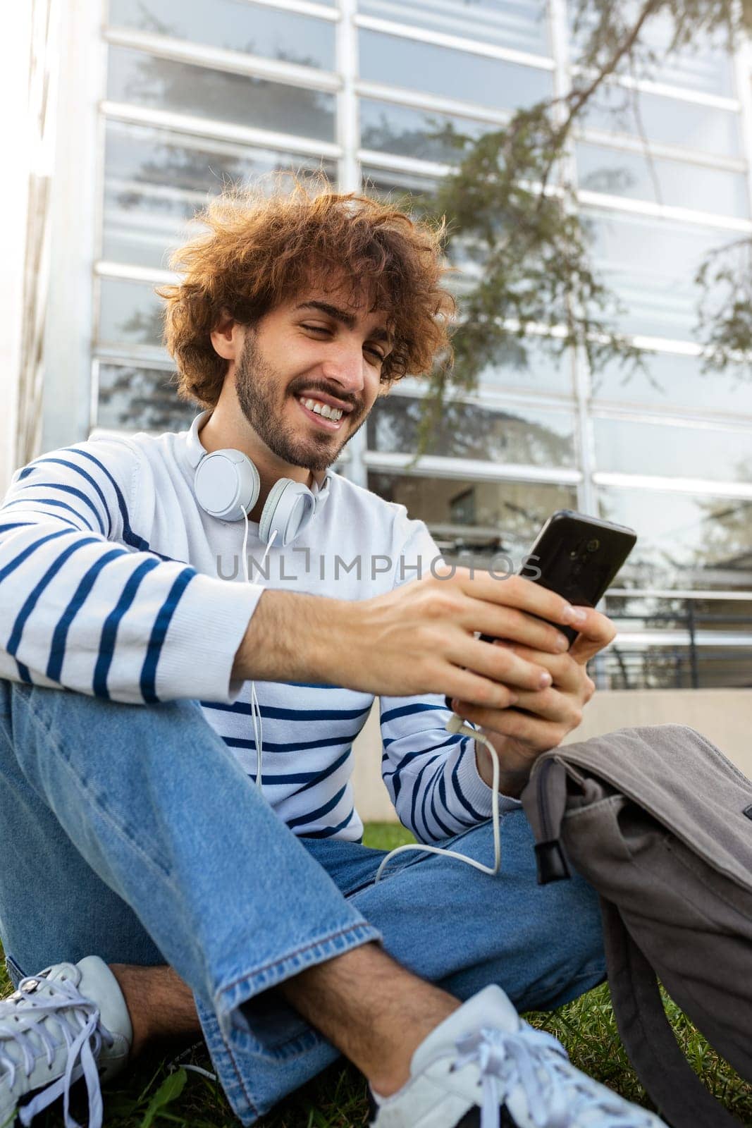Happy young male college student sitting on grass in campus using mobile phone. Vertical image. Education concept.