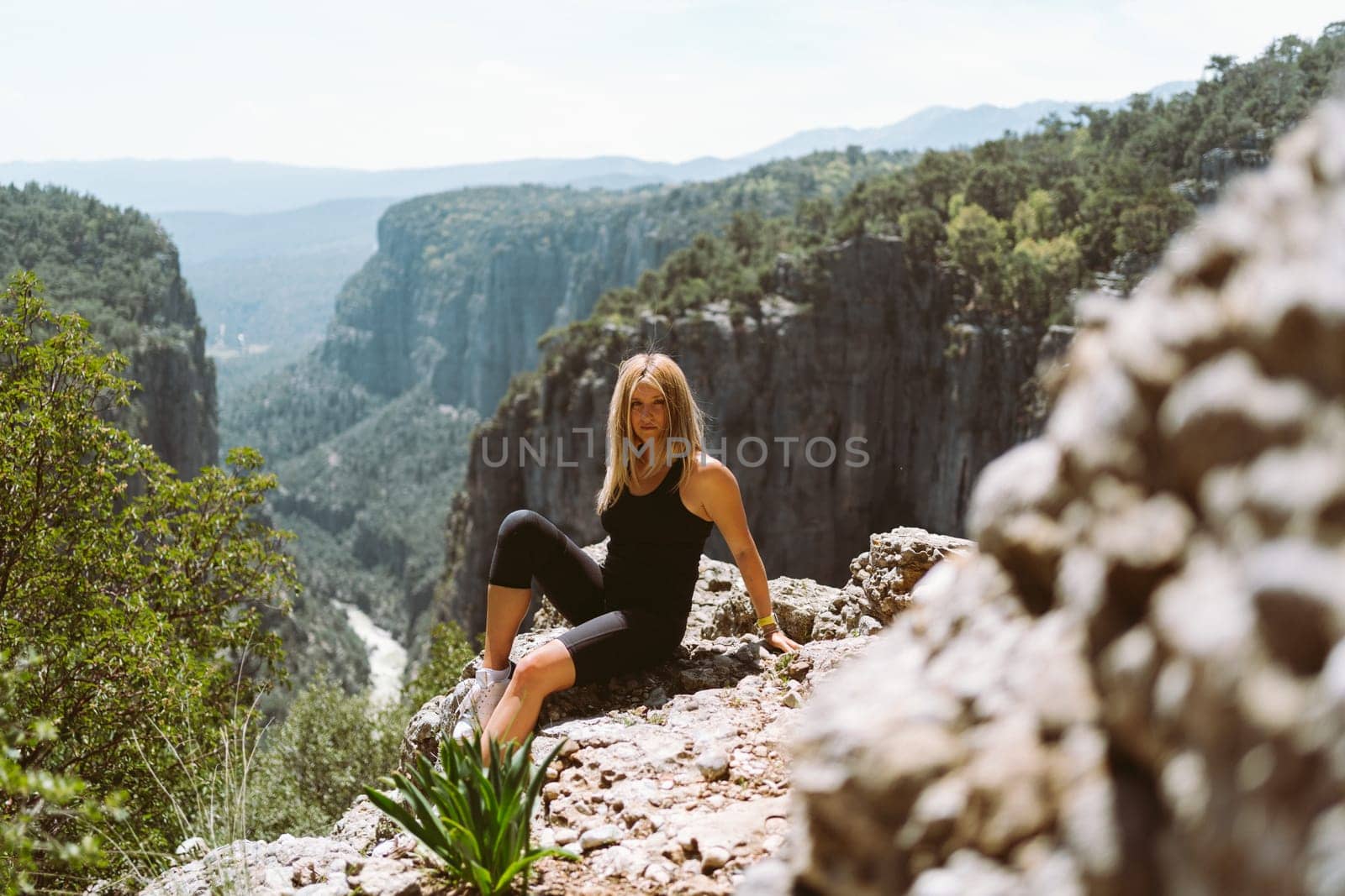 Young beautiful girl woman in black sporty slim outfit sitting on edge rock mountain cliff peak with view on valley canyon. Picture of fit female hiker reached the mountain top.