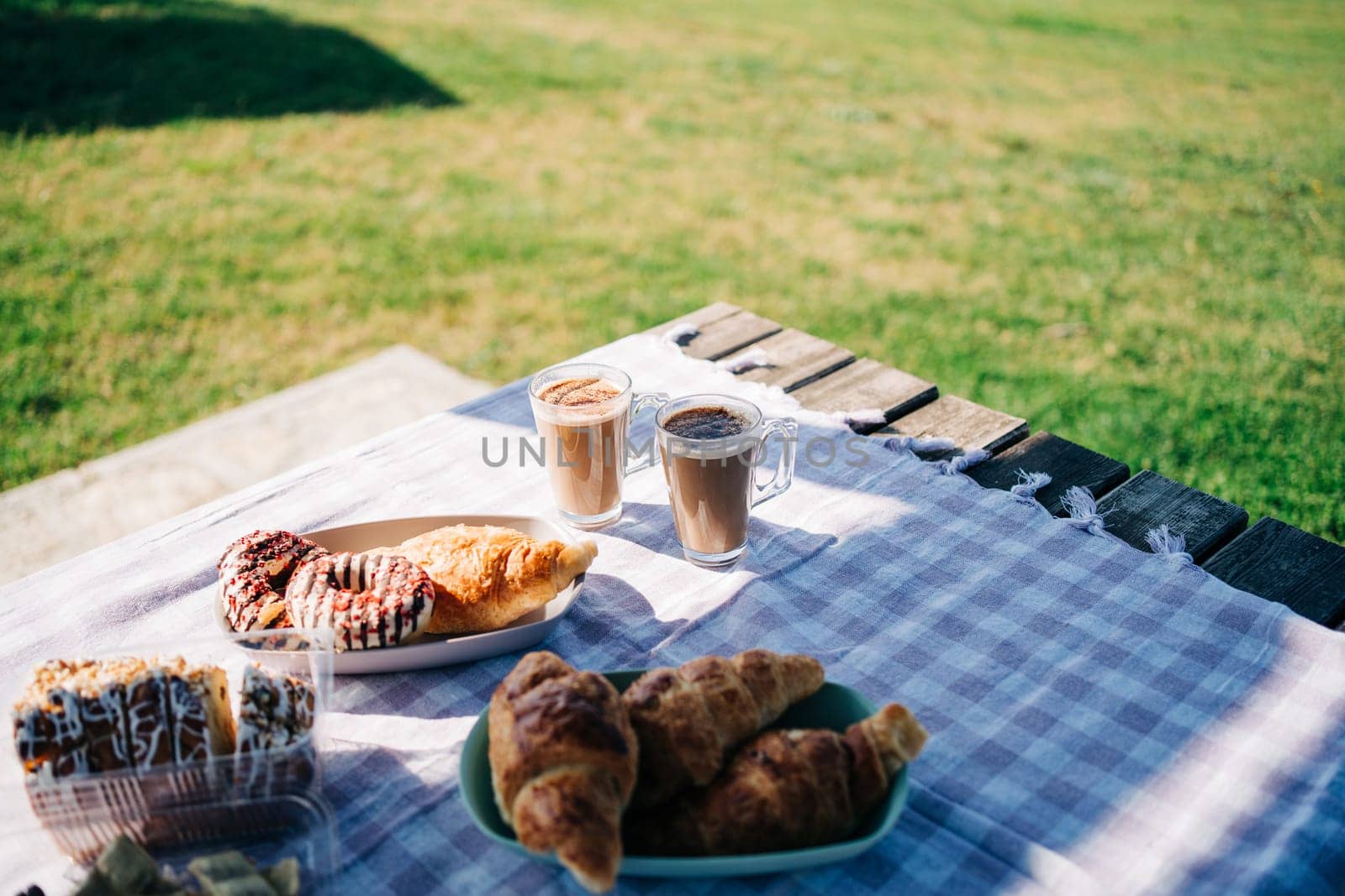 A picnic at the green lawn in the park. Closeup picture of breakfast with coffee, croissants and donuts on green grass outdoors meadow. by Ostanina