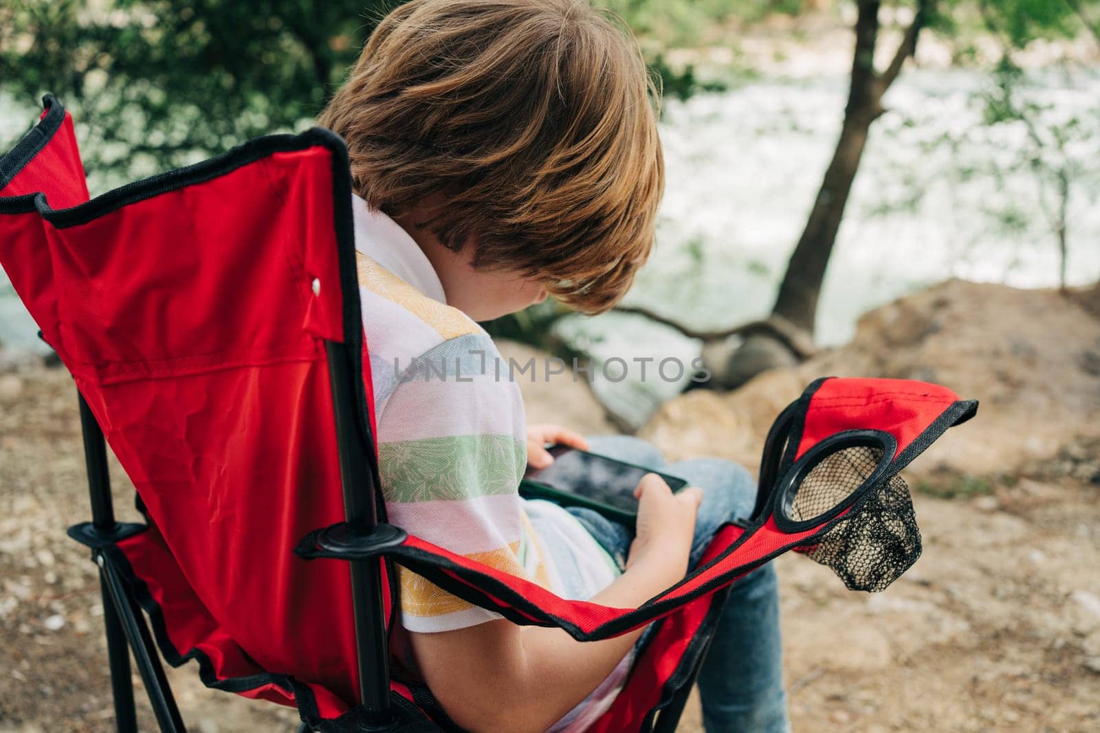 School boy kid child playing texting with his cellphone mobile smartphone while sitting on the red hiking foldable chair in the park outside outdoors