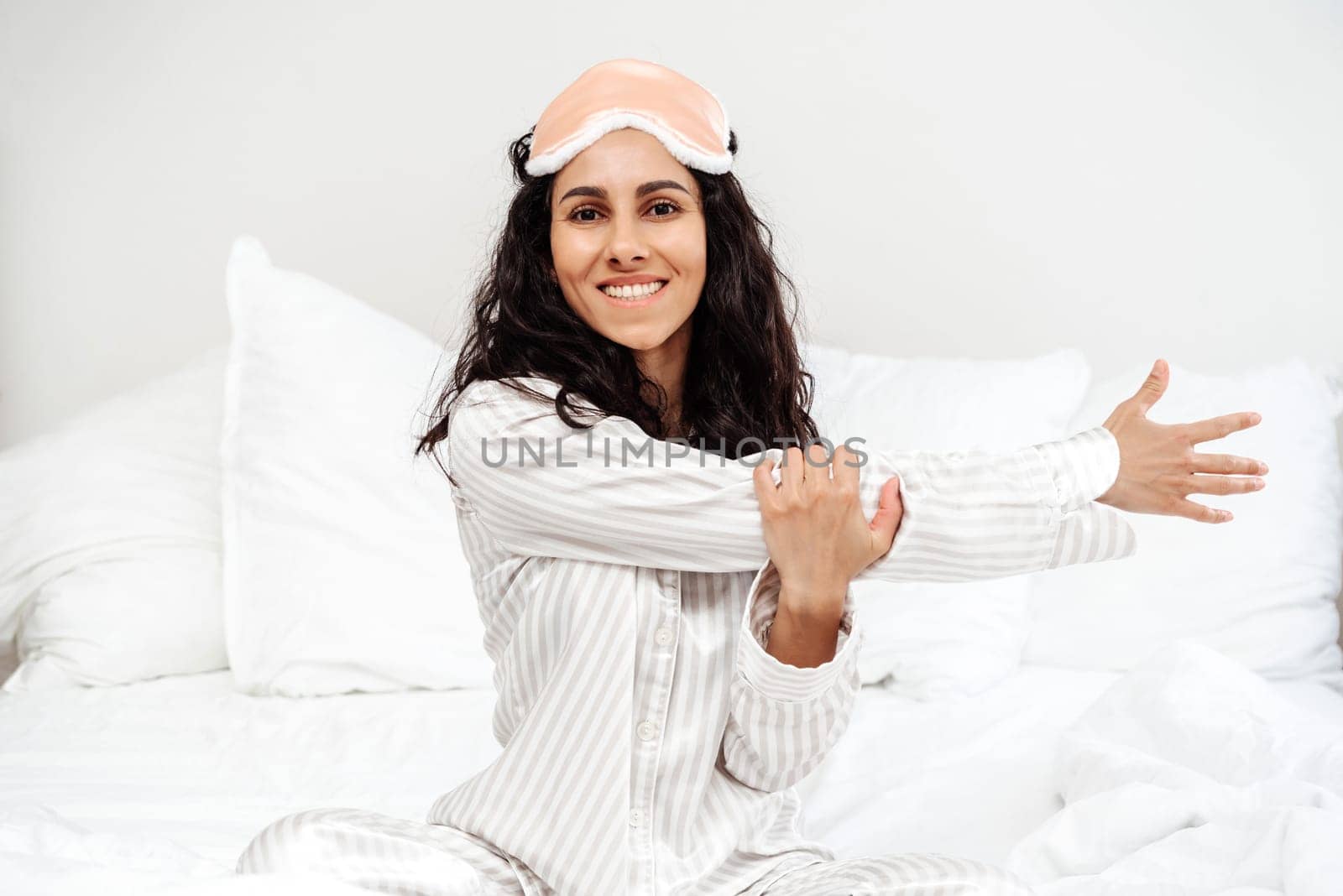 Young woman of Arab goes in for sports sitting in bed. The girl just woke up and kneads her shoulders and arms after a healthy sleep. She smiles brightly and feels a surge of energy and positive