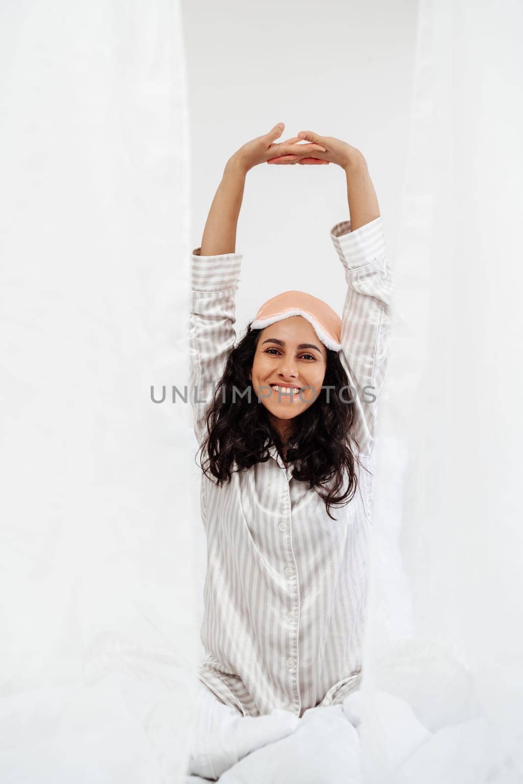 Young positive Hispanic woman sitting on bed after waking up in morning, stretching arms up as a morning ritual and exercise for health. Beautiful girl in pajamas in a snow-white room.