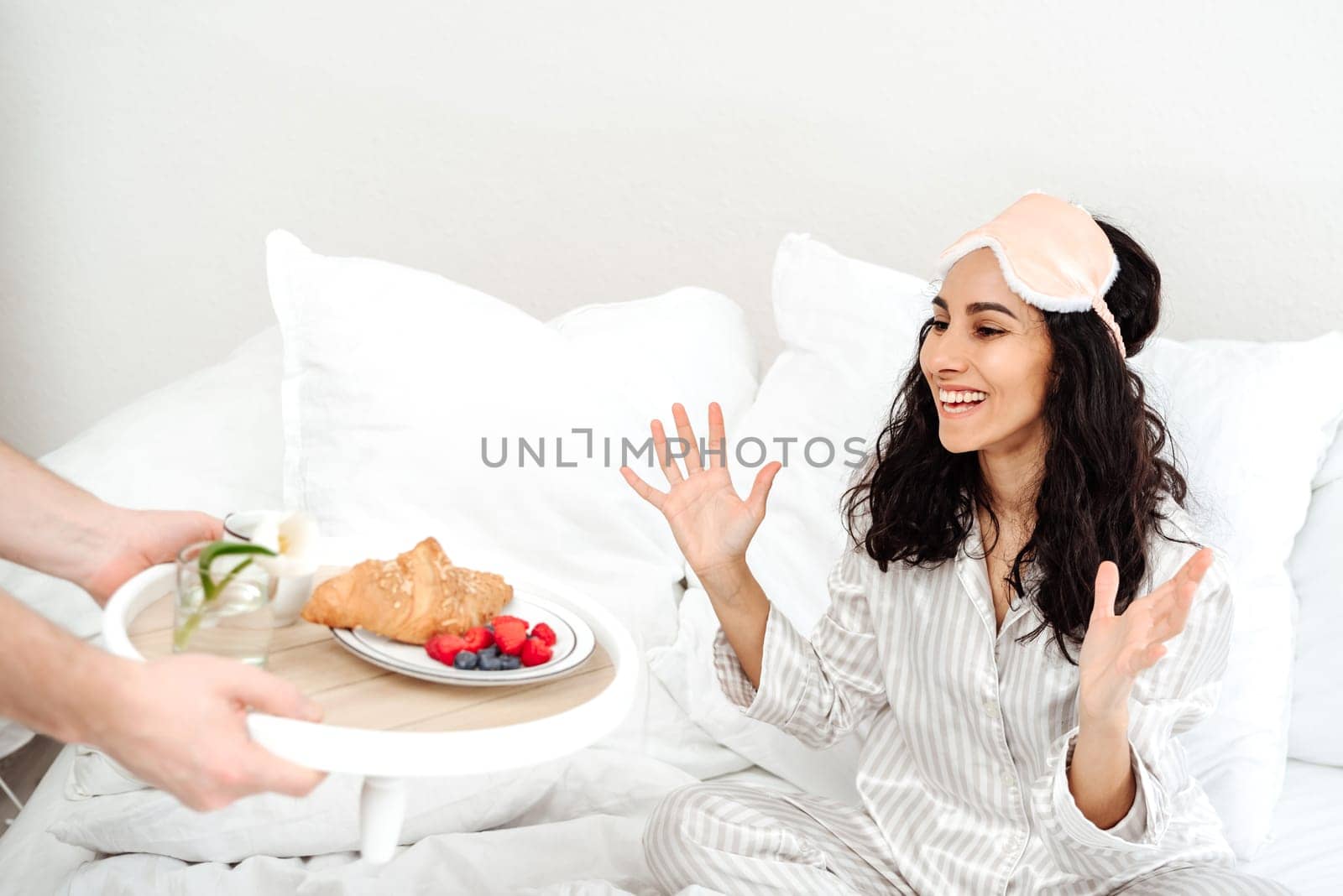 Beautiful woman of Eastern appearance rejoices in breakfast in bed. Hands holding a tray with croissant, berries and a flower. Girl in pajamas has just woken up and brightly rejoices at the surprise.