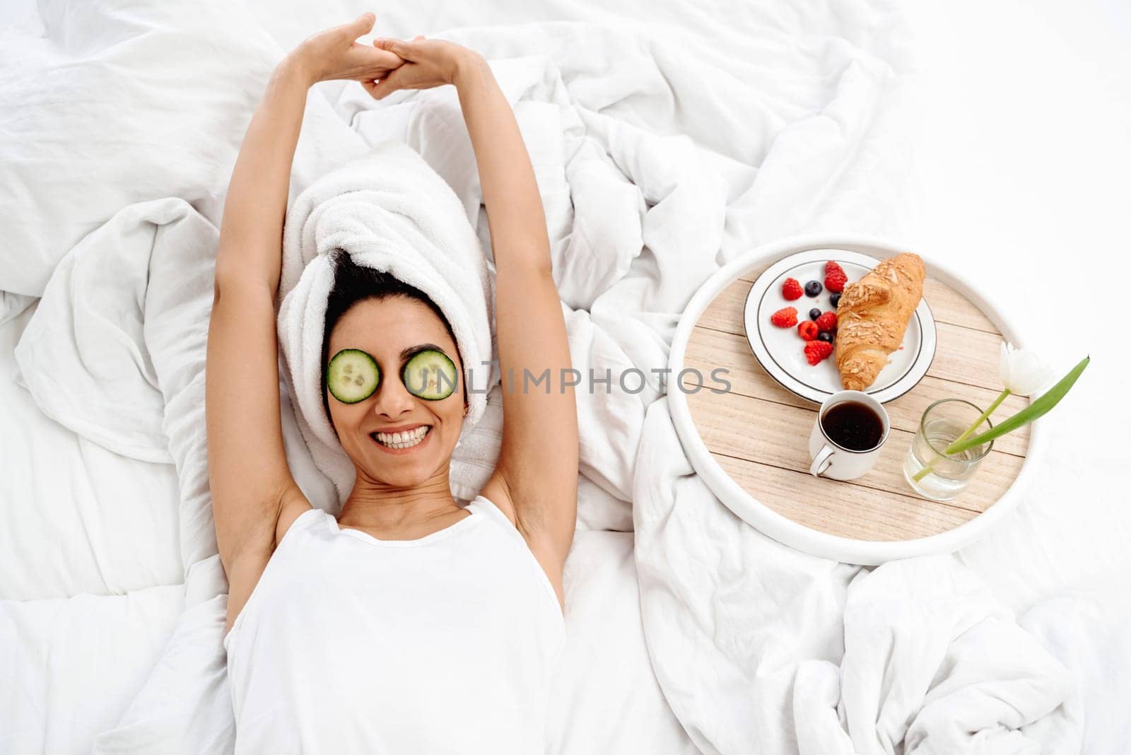Happy woman stretches in bed in the morning with a breakfast tray. She just got out of the shower and put cucumber slices on her eyes as a facial. The girl lies on a snow-white bed, view from above