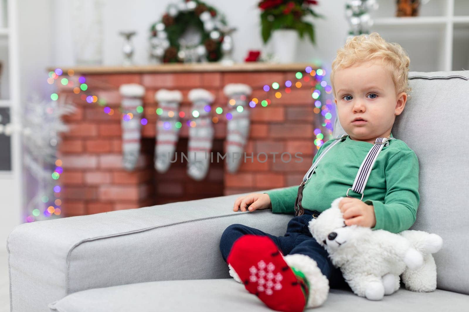 A little boy with curly blond hair sits on a gray couch. The child holds a white cuddly toy in his hand. A brick fireplace can be seen in the background. The fireplace is decorated with colorful Christmas lights.