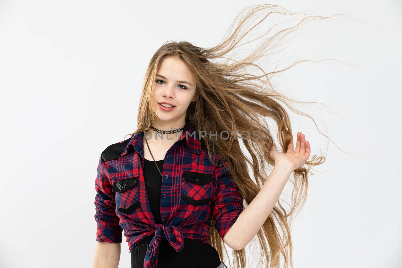 A girl in a youthful outfit poses for a photo against a white background. She is combing her fingers through her long, flowing hair. The woman is wearing a red and blue checked shirt.
