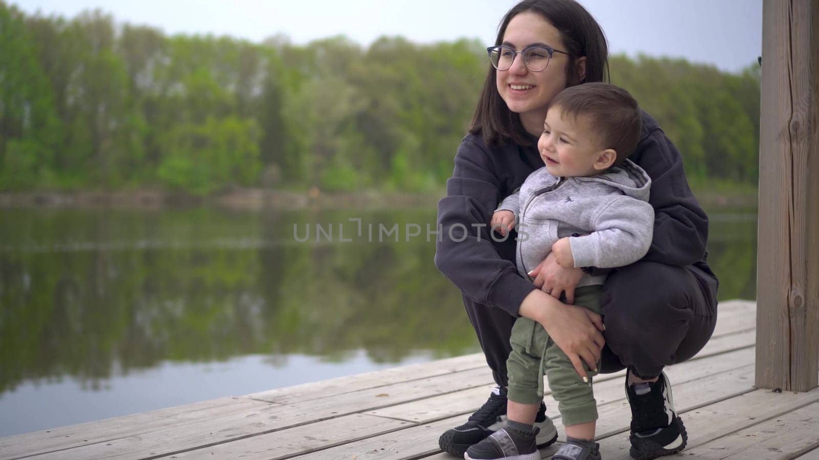 A young mother stands with her son on a pier by the river. Woman with a child in nature. 4k