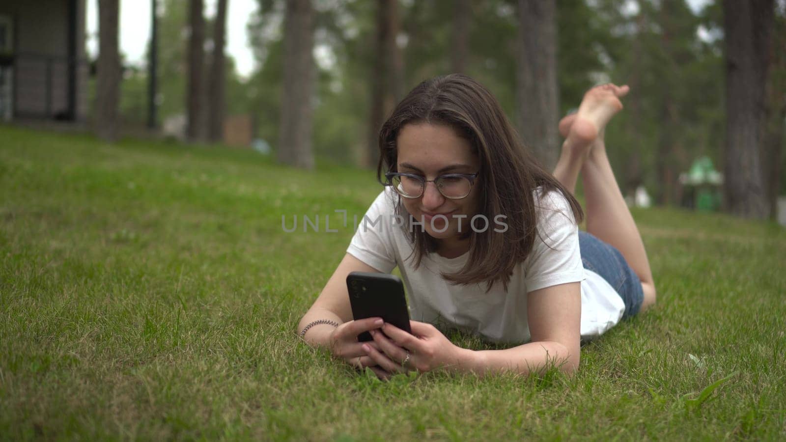 A young woman lies on the grass and chats on a smartphone. A girl with glasses is resting in the park with a phone. 4k