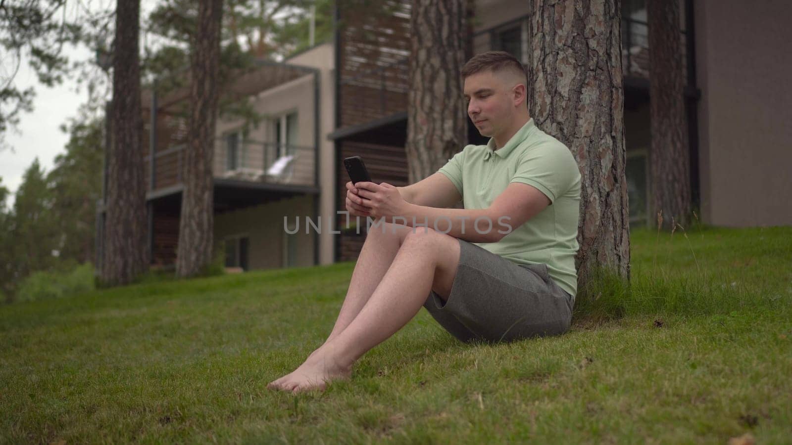 A young man sits under a tree near a country house with a phone in his hands. A man is texting on a smartphone in nature on the grass. 4k
