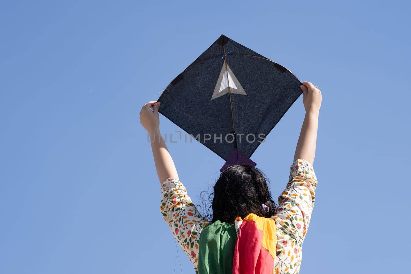 Young girl in traditional indian clothing holding black kite high above head launching it on sankranti republic independence day celebrations by Shalinimathur