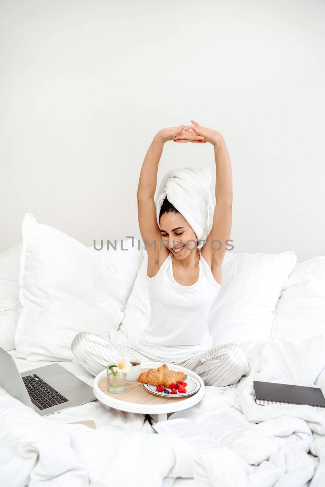Happy woman stretching in bed in the morning with breakfast tray. The girl is ready to start work and perform tasks after a delicious healthy breakfast. A towel is wrapped around the woman's head.