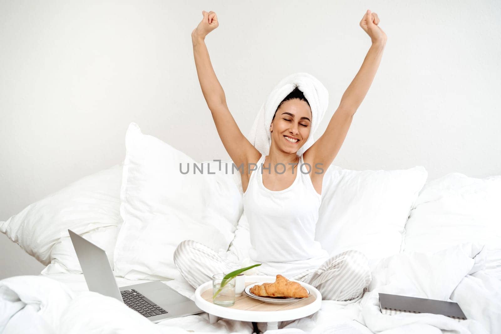 Beautiful young latin woman stretching on a bed in the morning, warm sun shines on her from the big window. Uses laptop and eating croissants and coffee for breakfast. Work from home, stay home concept.