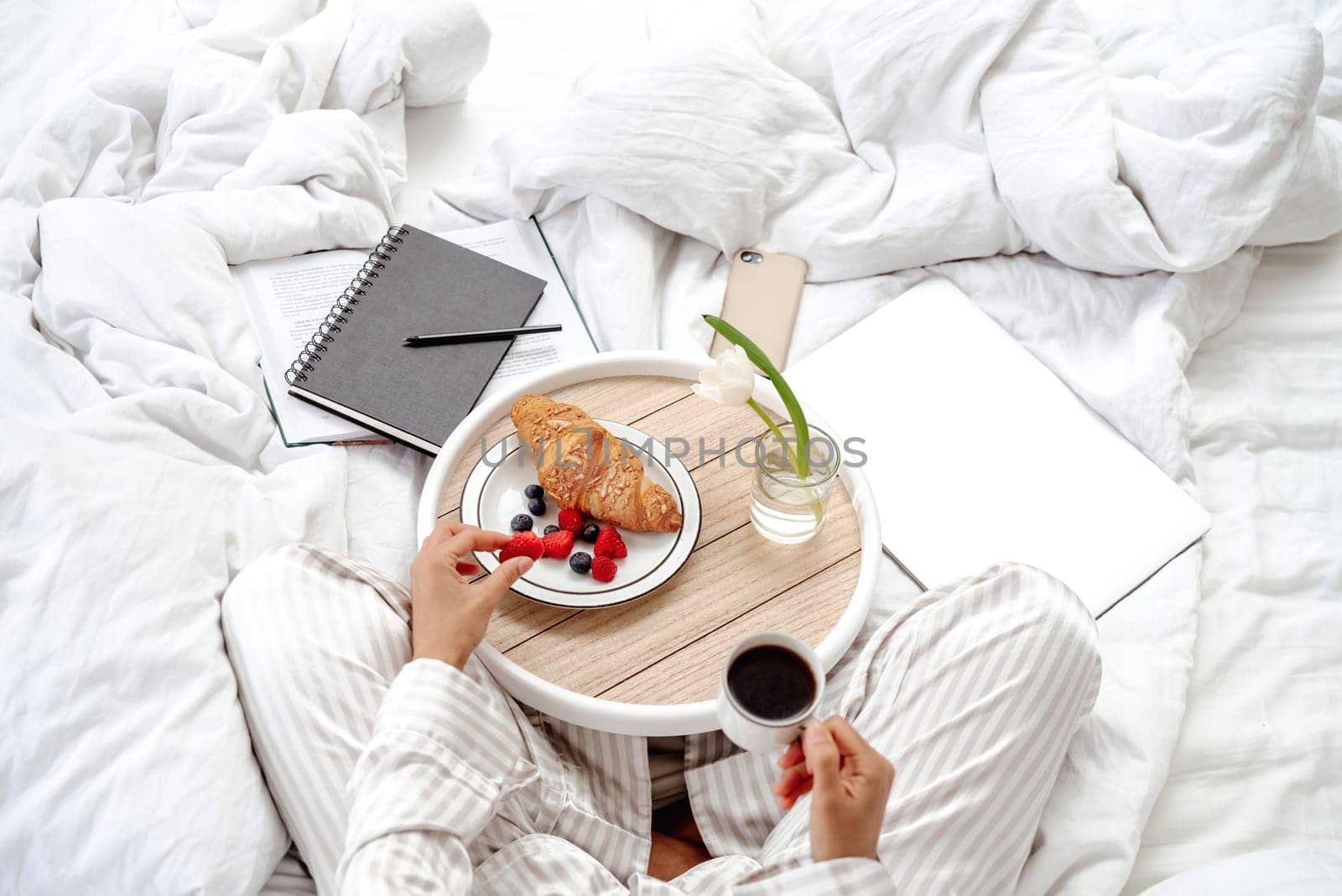 Healthy breakfast in bed croissant and fresh berries. The girl holds a cup of coffee in her hand and is ready to enjoy the meal. Starting a productive day in bed with work supplies laptop and notebook