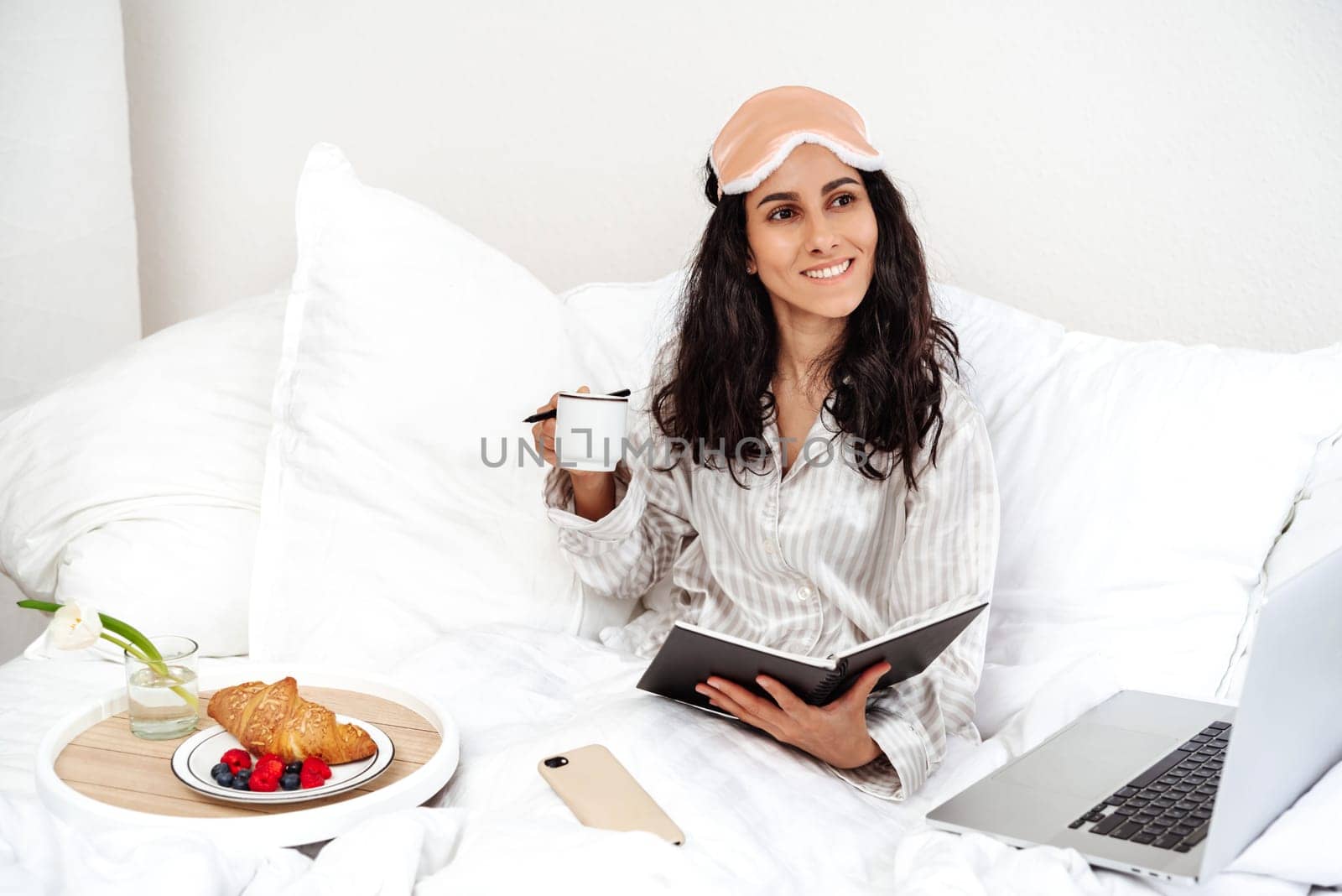 Beautiful oriental millennial woman having breakfast in bed. The girl plans things and deals with working moments in the process rest. The girl just woke up and took off the mask from her eyes