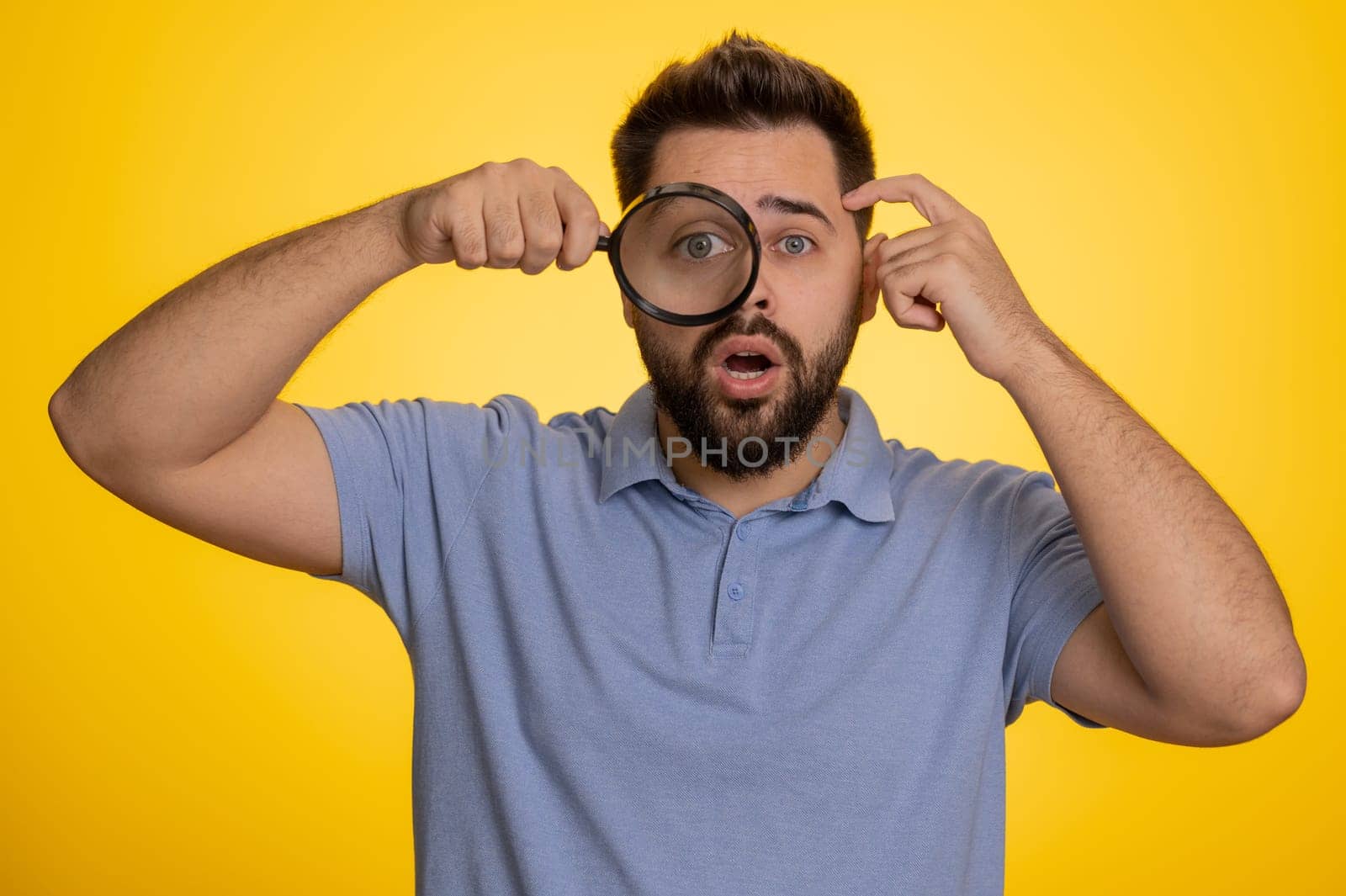Investigator researcher scientist man holding magnifying glass near face looking into camera with big zoomed funny eyes, searching analysing. Excited caucasian guy isolated on yellow studio background