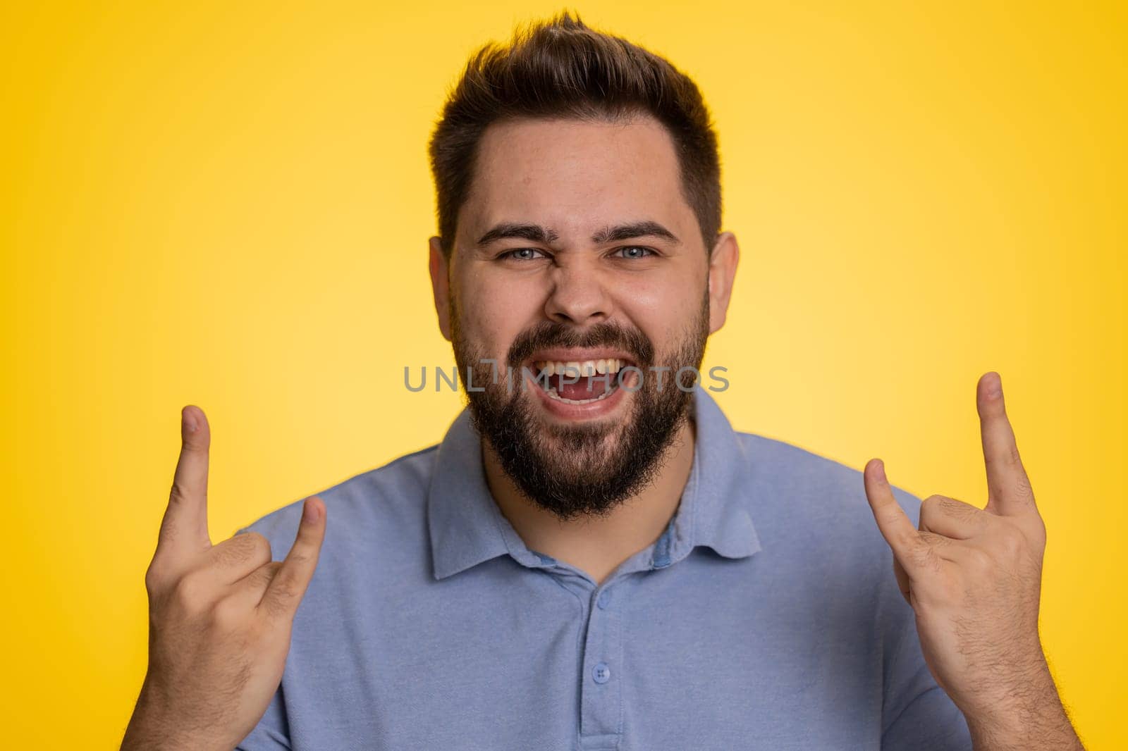 Overjoyed caucasian man showing rock n roll gesture by hands, cool sign, shouting yeah with crazy expression, dancing, emotionally rejoicing in success. Young happy guy isolated on yellow background