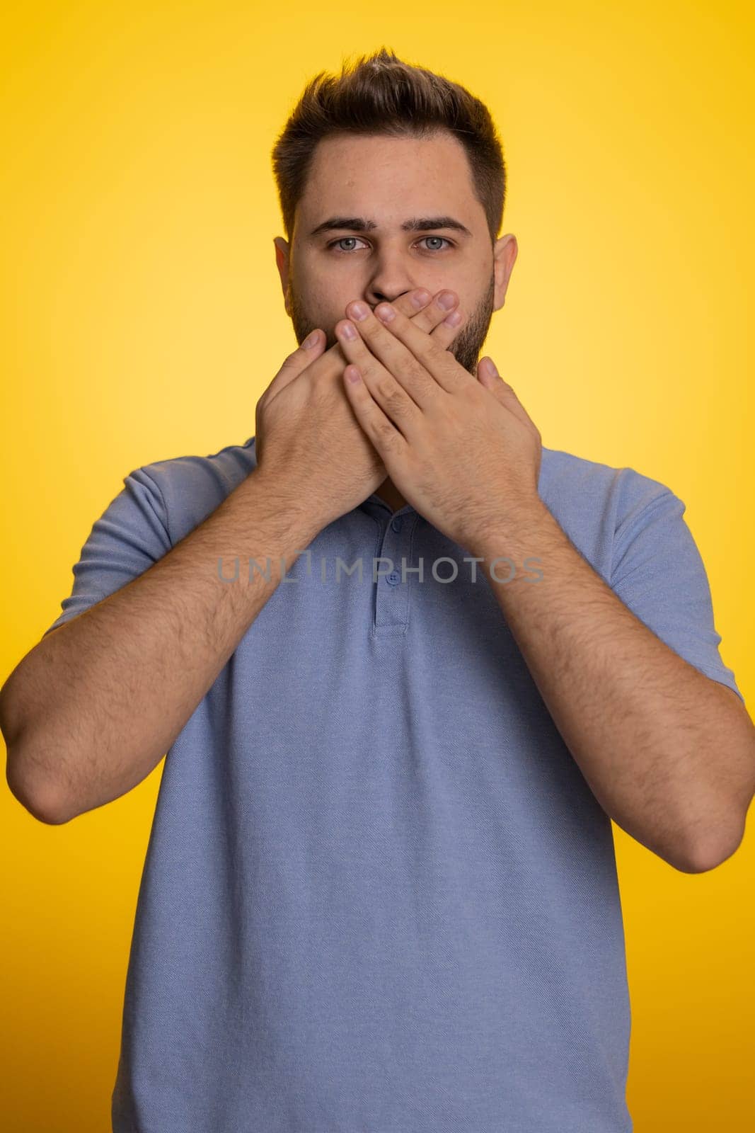 I will not say anyone. Frightened man closing his mouth with hand, looking intimidated scared at camera, gestures no, refusing to tell terrible secret, unbelievable truth. Guy on yellow background