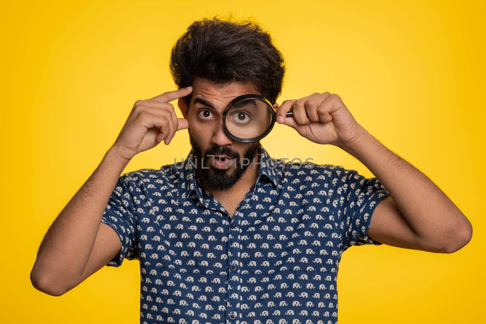 Investigator researcher scientist man holding magnifying glass near face, looking into camera with big zoomed funny eyes, searching, analysing. Handsome indian guy isolated on yellow studio background