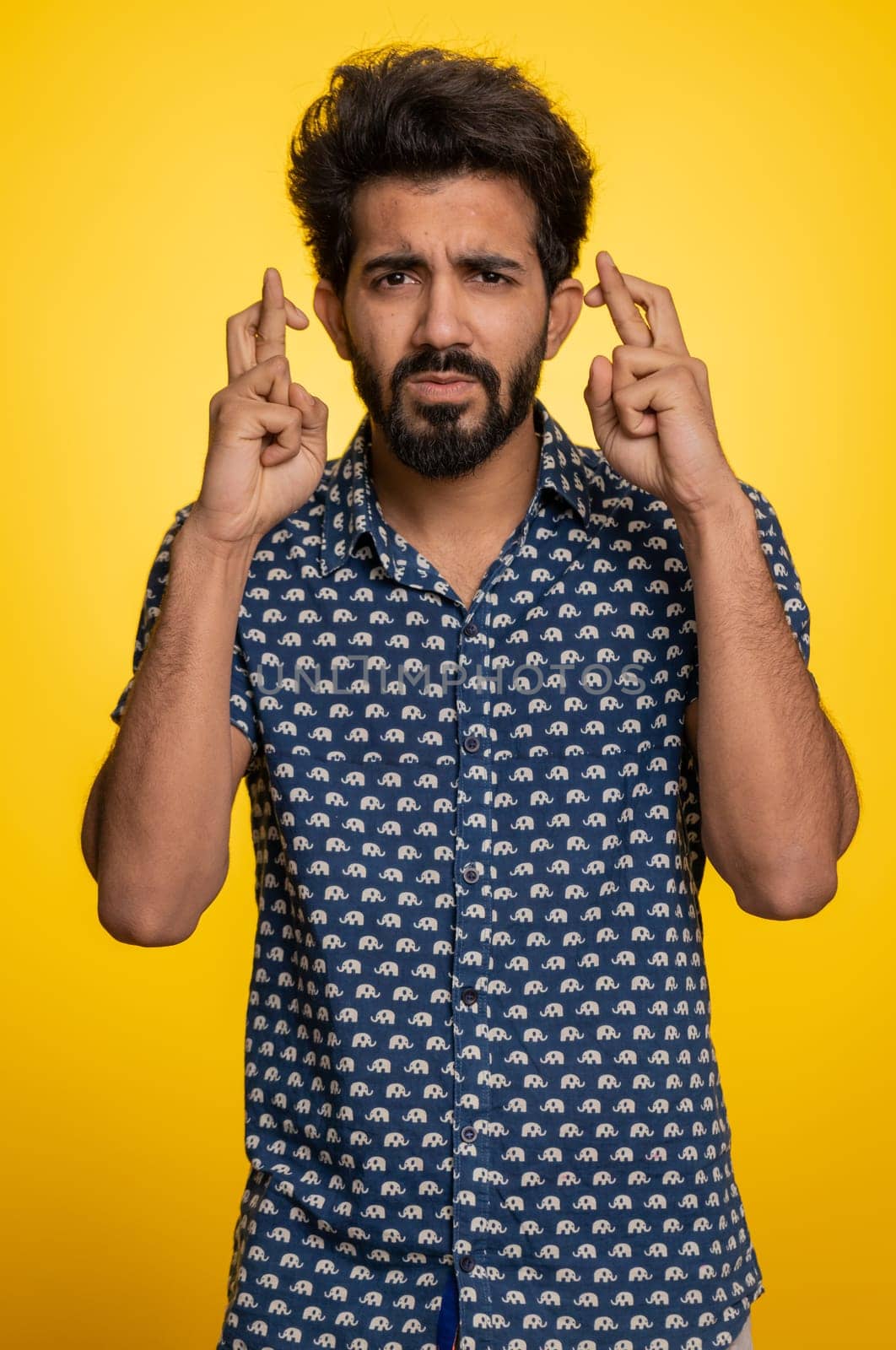 Indian young man crossed fingers looking at camera asking for good lucky news, wishing good exam results, dreaming about win victory in lottery jackpot. Hindu guy isolated on yellow studio background