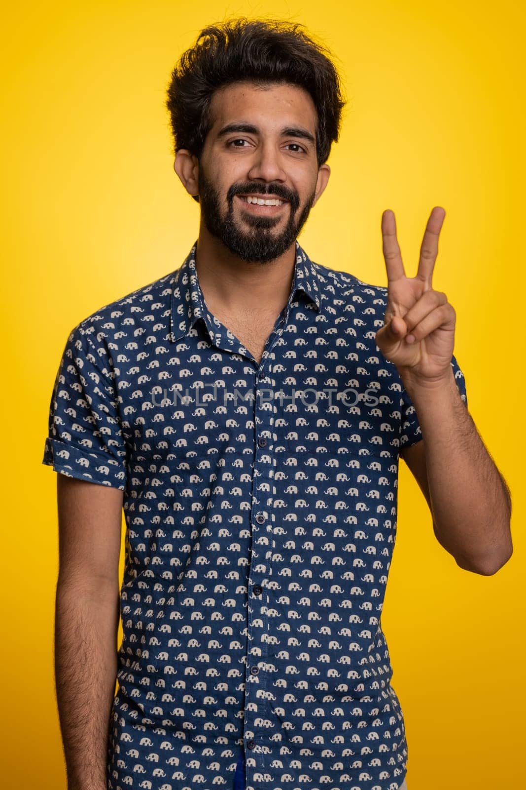 Young indian man showing victory sign, hoping for success and win, doing peace gesture, smiling with kind optimistic expression. Handsome bearded hindu guy isolated alone on yellow studio background