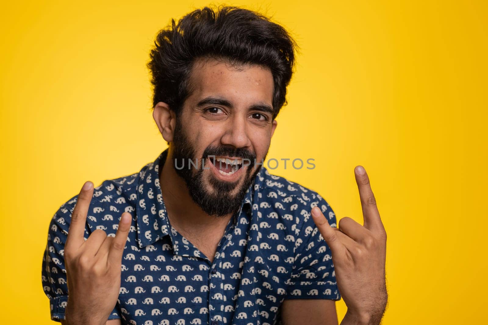 Overjoyed young indian man showing rock n roll gesture by hands, cool sign, shouting yeah with crazy expression, dancing, emotionally rejoicing in success. Hindu guy isolated on yellow background