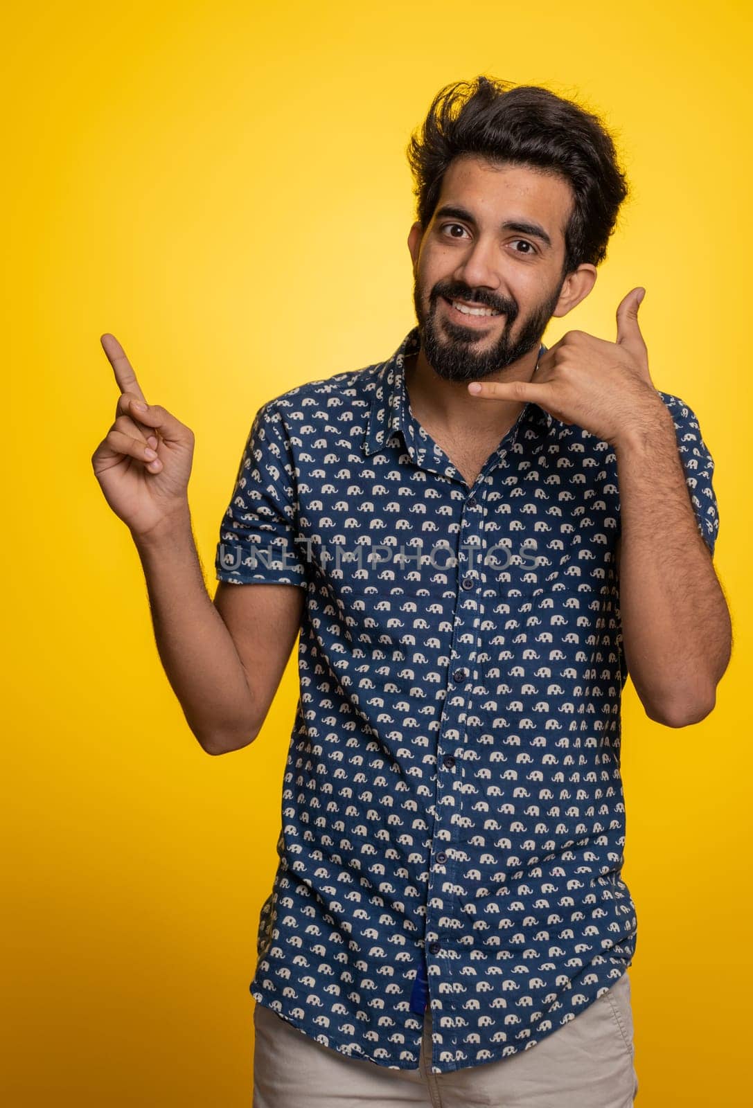 Call us, here is contact number. Handsome bearded indian man in shirt looking at camera doing phone gesture like says hey you call me back conversation. Hindu guy isolated on yellow background, indoor