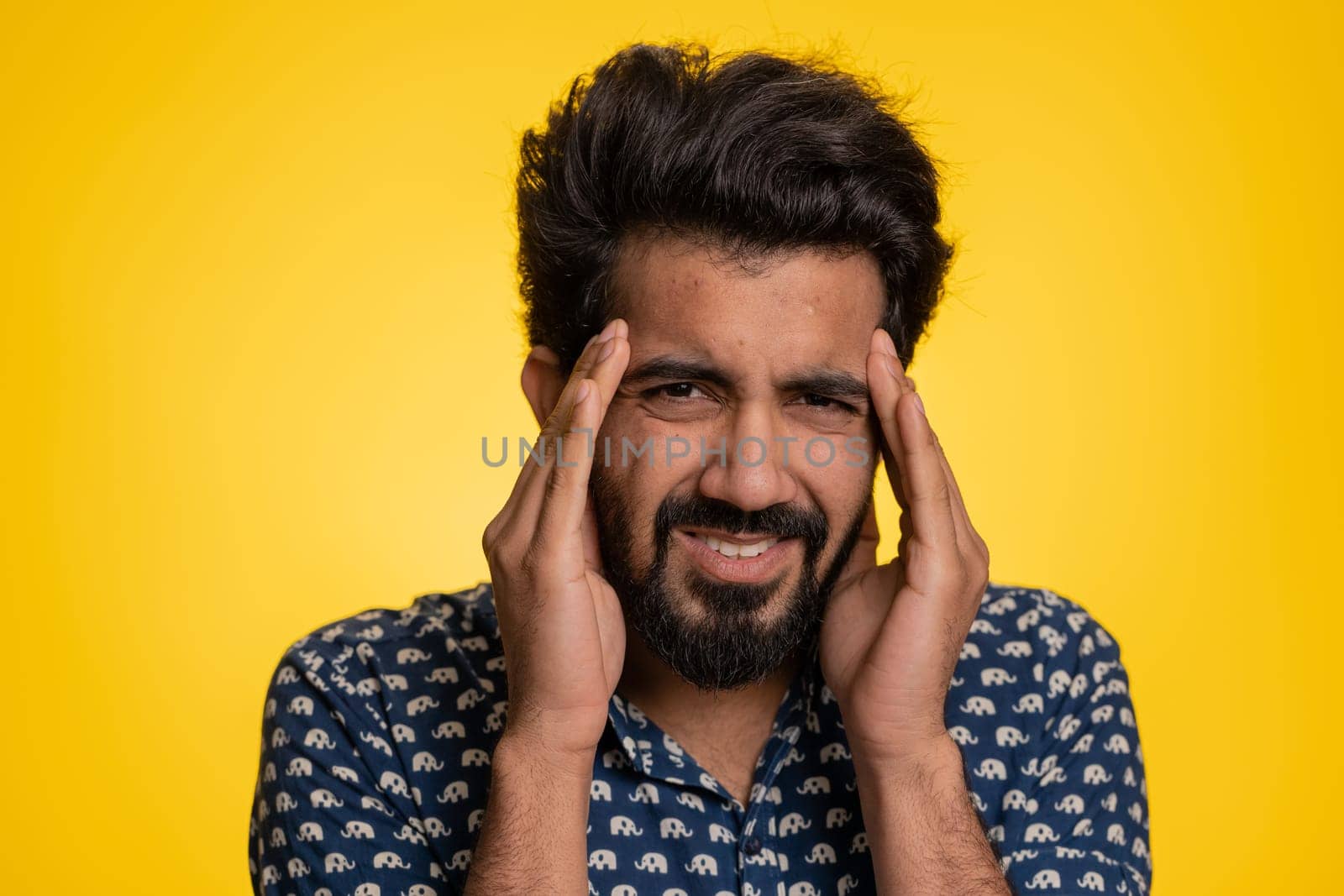 Handsome indian bearded man in shirt rubbing temples to cure headache problem, suffering from tension, migaine, stress, grimacing in pain, high blood pressure isolated alone on yellow wall background