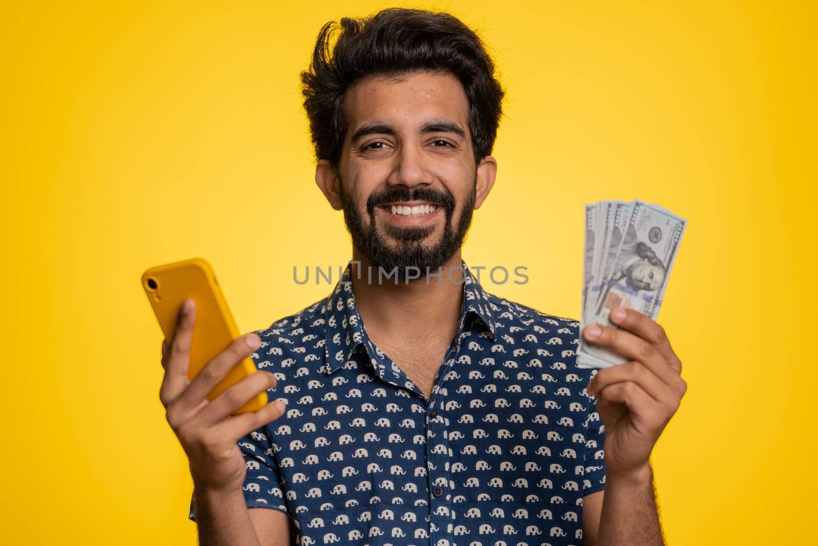 Indian man looking smartphone display sincerely rejoicing win, receiving money dollar cash banknotes, success lottery jackpot luck, income salary wage. Hindu guy isolated on studio yellow background