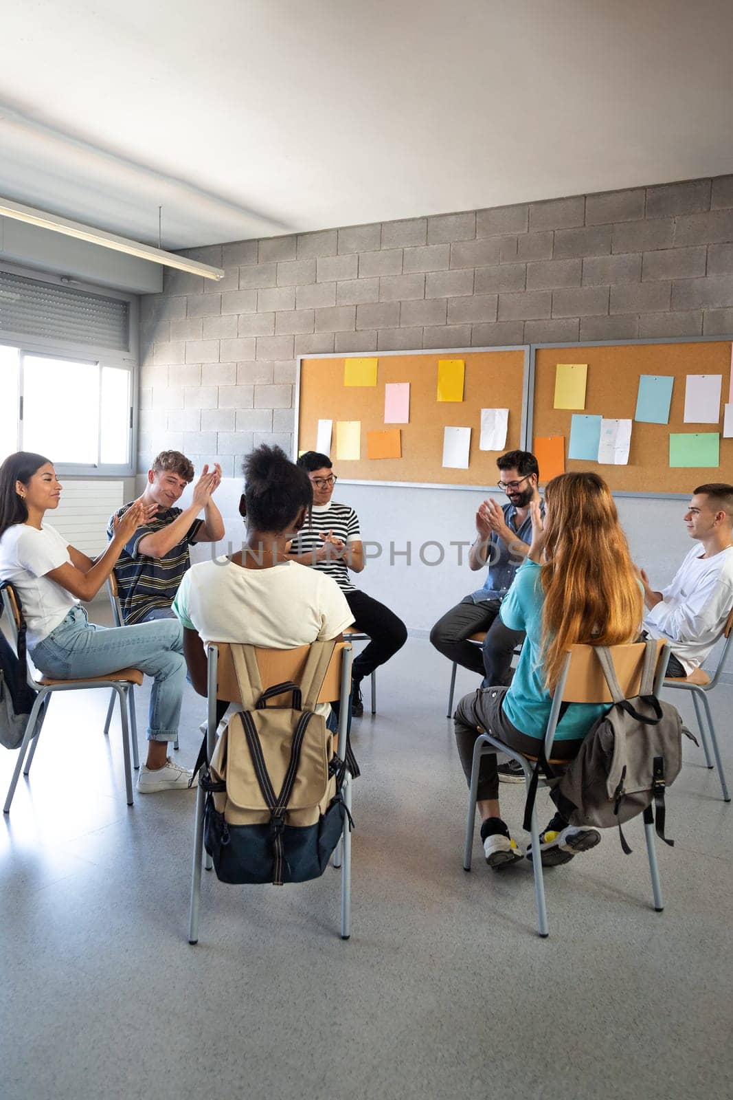 Multiracial high school students sitting in a circle clapping together celebrating achievement. Support group. Vertical. by Hoverstock