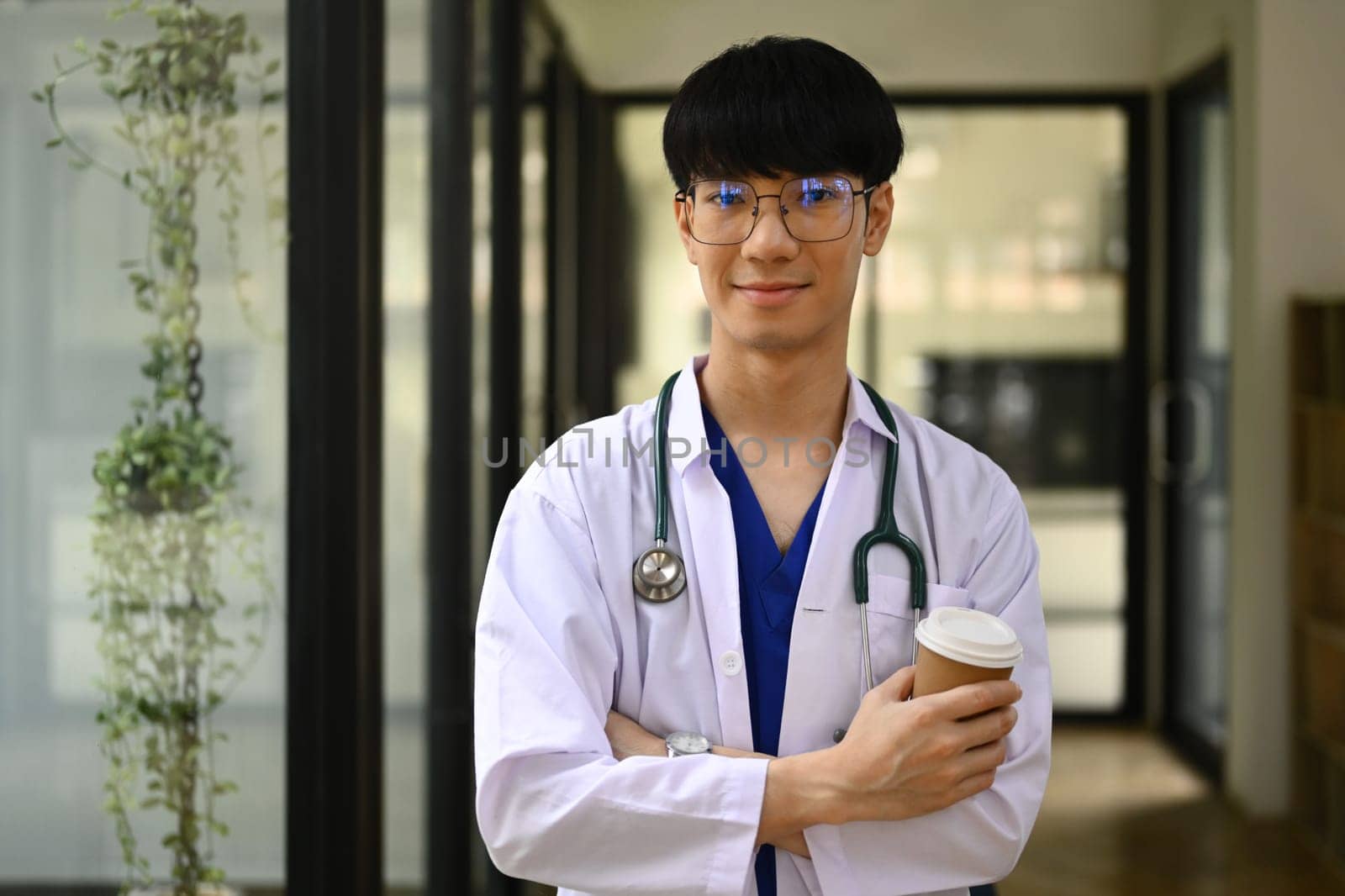 Smiling general practitioner in white coat with stethoscope standing at hospital hallway and smiling to camera.