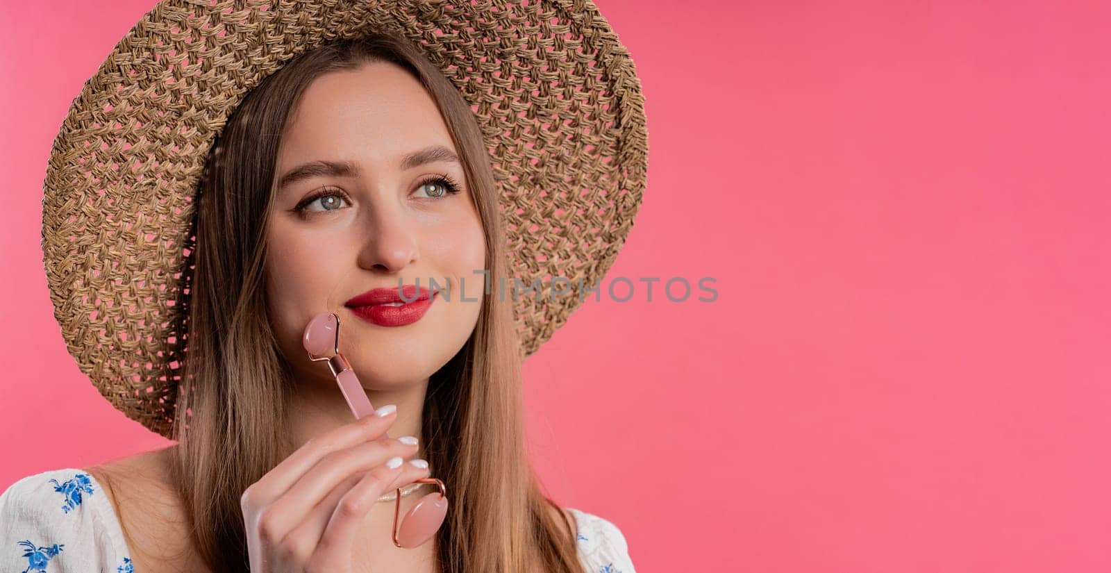 Blonde woman doing face massage with rose quartz stone roller on pink background. Facial self care, beauty rituals, cosmetology, anti aging and anti-wrinkle treatment