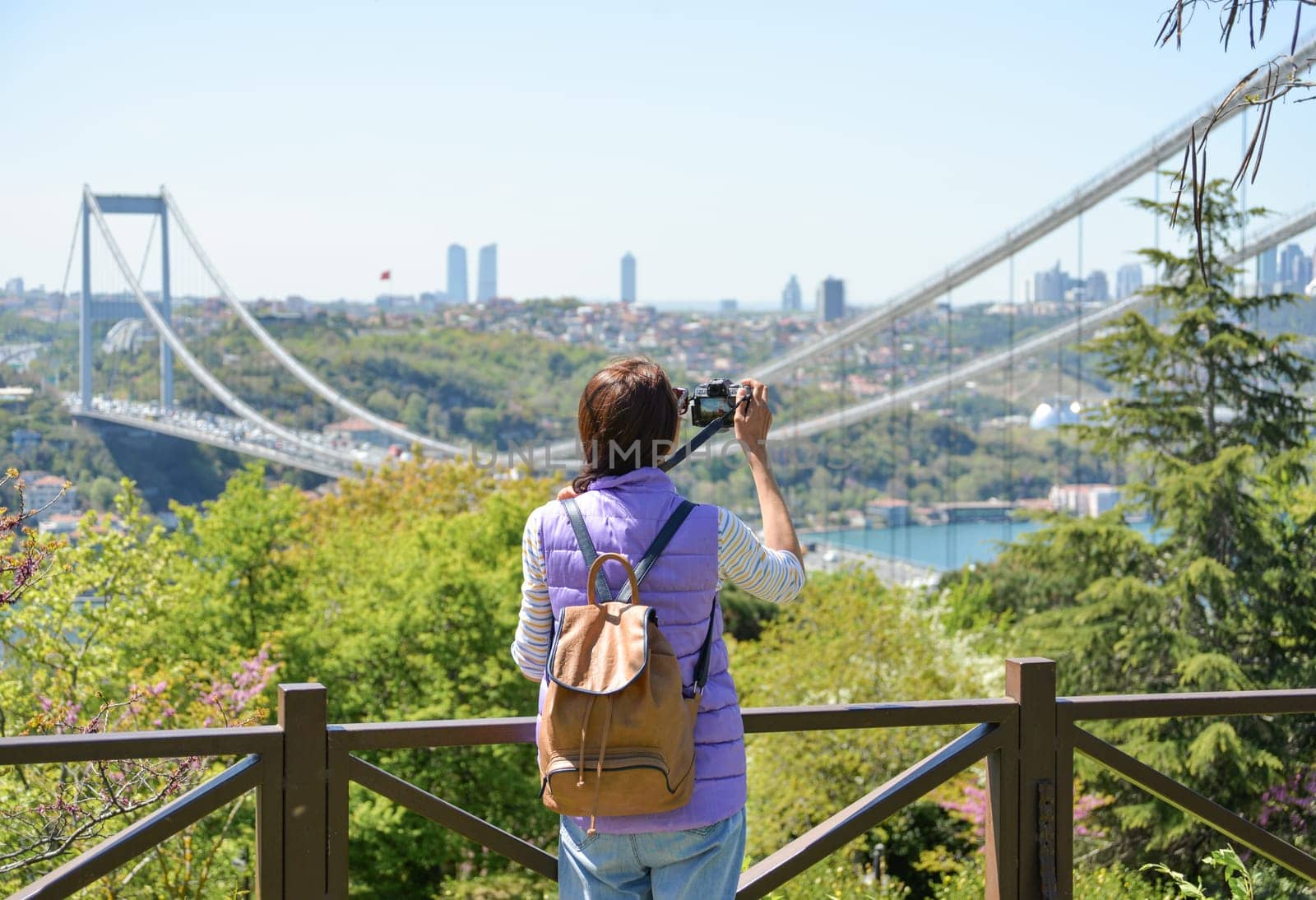 A young woman tourist with a backpack photographs the cityscape with a panoramic view of the Fatih Sultan Mehmet Bridge. Back view. Otagtepe Park.