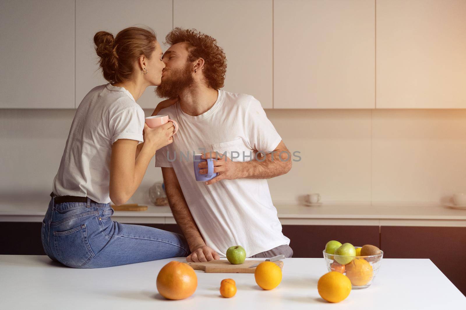 Kissing beautiful couple embracing each other enjoying their their morning coffee in a new house. Making fun at modern kitchen and kissing while cooking at home. New house concept by LipikStockMedia