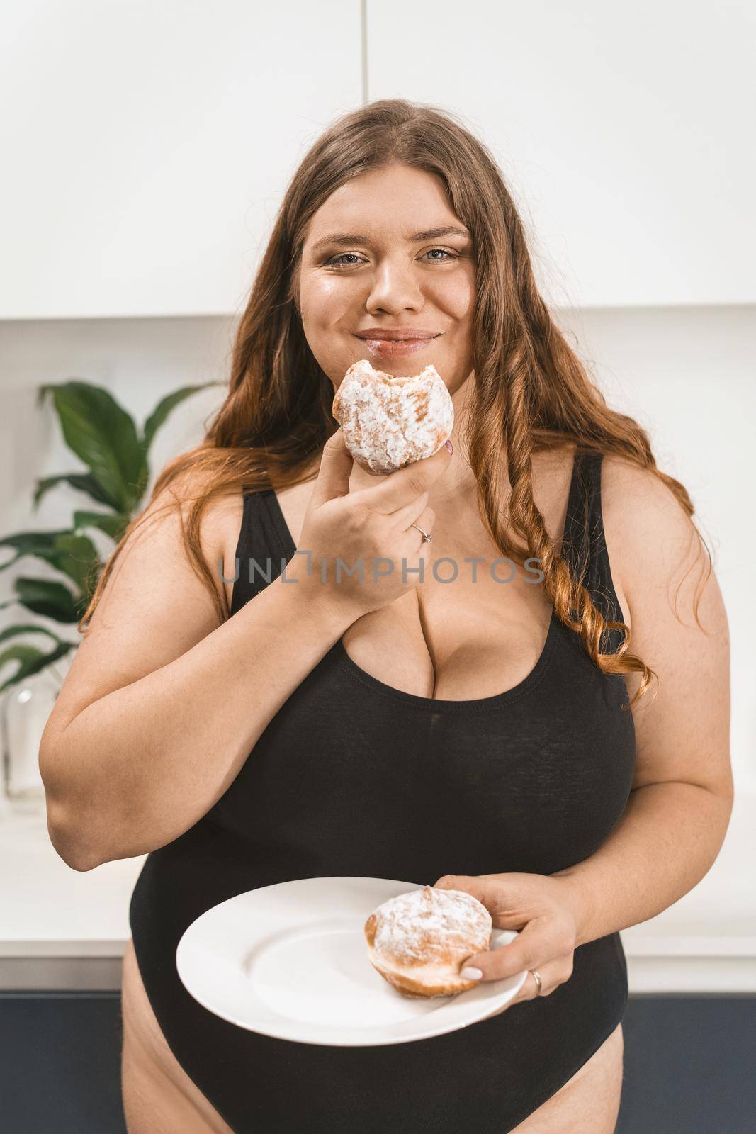 Satisfied young fat woman eating a cake happy smiling on camera wearing black swimsuit. Beautiful chubby young woman eating unhealthy food. Fat girl eating cake standing on modern kitchen.