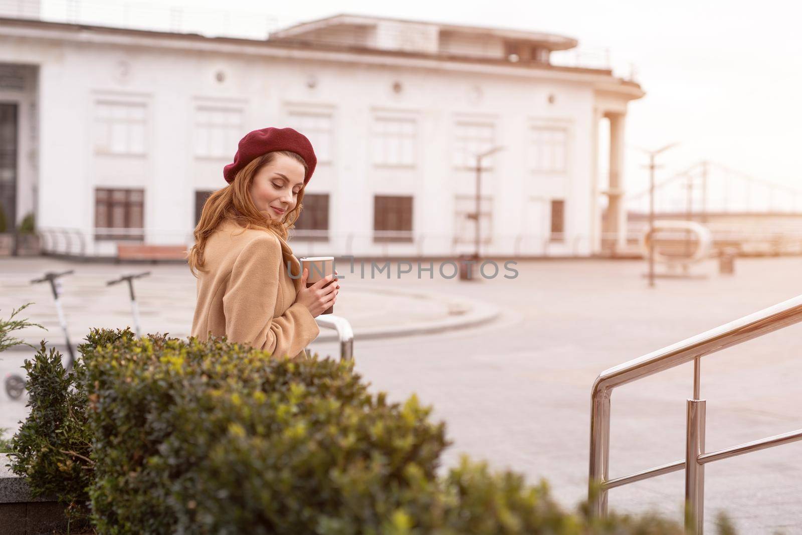 Tender beautiful french girl in an autumn beige coat standing at flower beds with share it or rent electric scooters on background waiting for her date holding coffee mug. Warm toned photo.