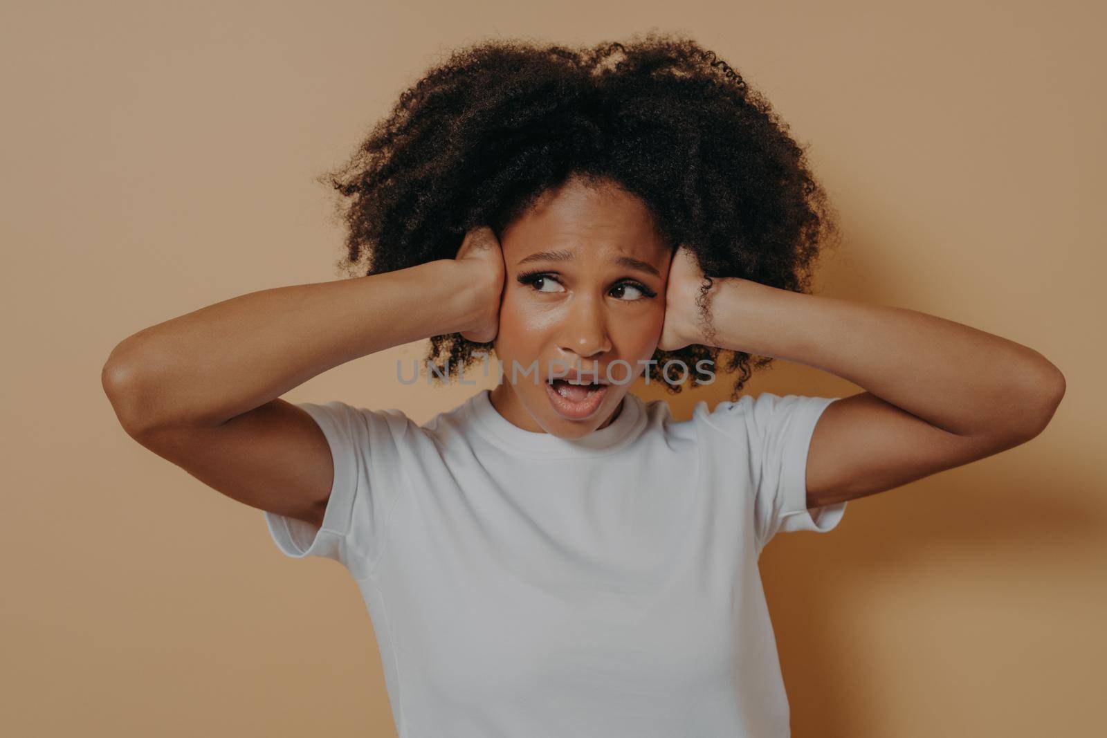 Young annoyed african woman covering her ears and keeping mouth opened isolated on beige background by vkstock