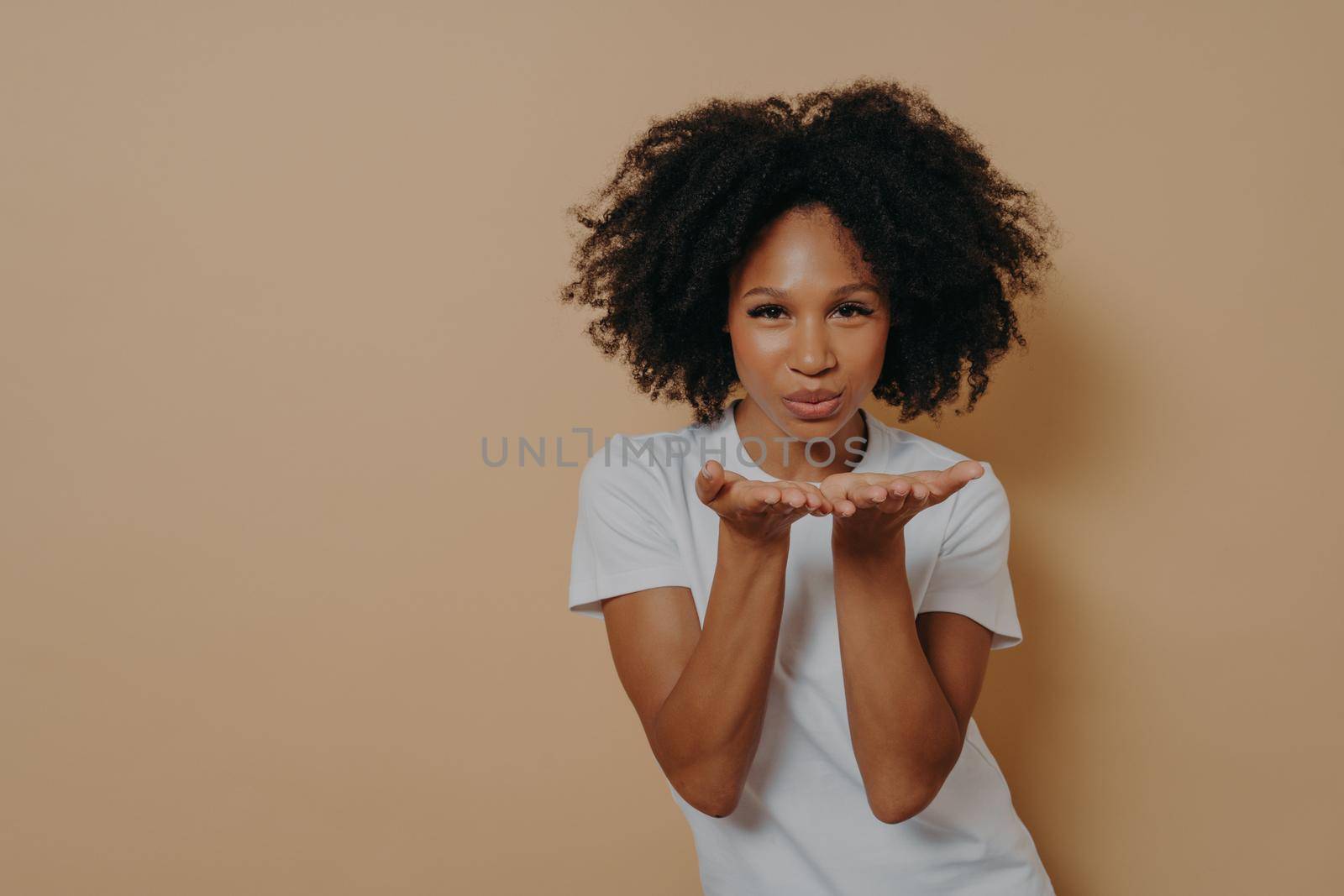 Beautiful curly flirty african woman in white t-shirt sending air kiss and smiling, demonstrating warm feelings and love while posing in studio on beige background with copy space for text