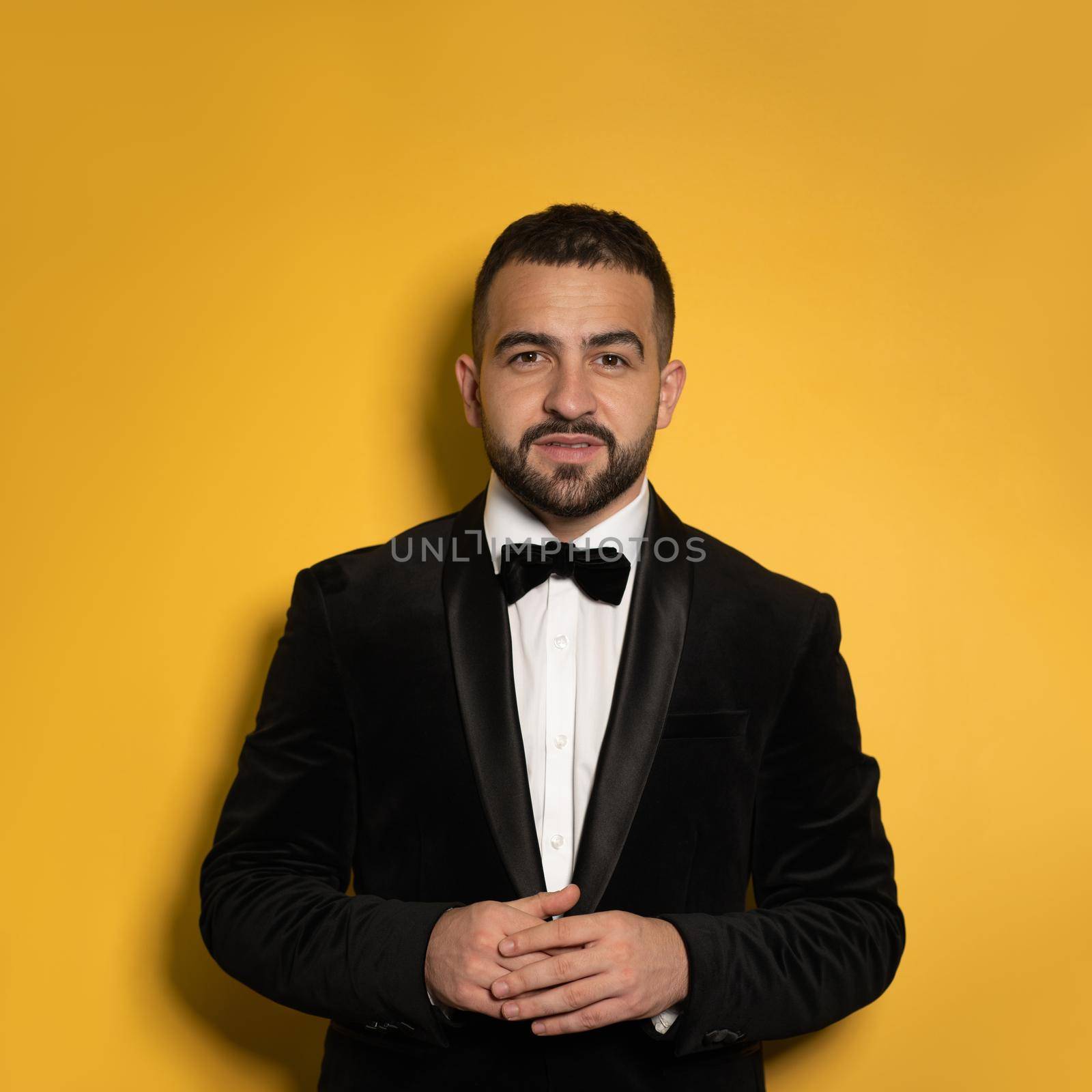 Concentrated handsome man wearing black tuxedo with hands folded looking on camera. Handsome young smiling caucasian man isolated on yellow background. Square crop.