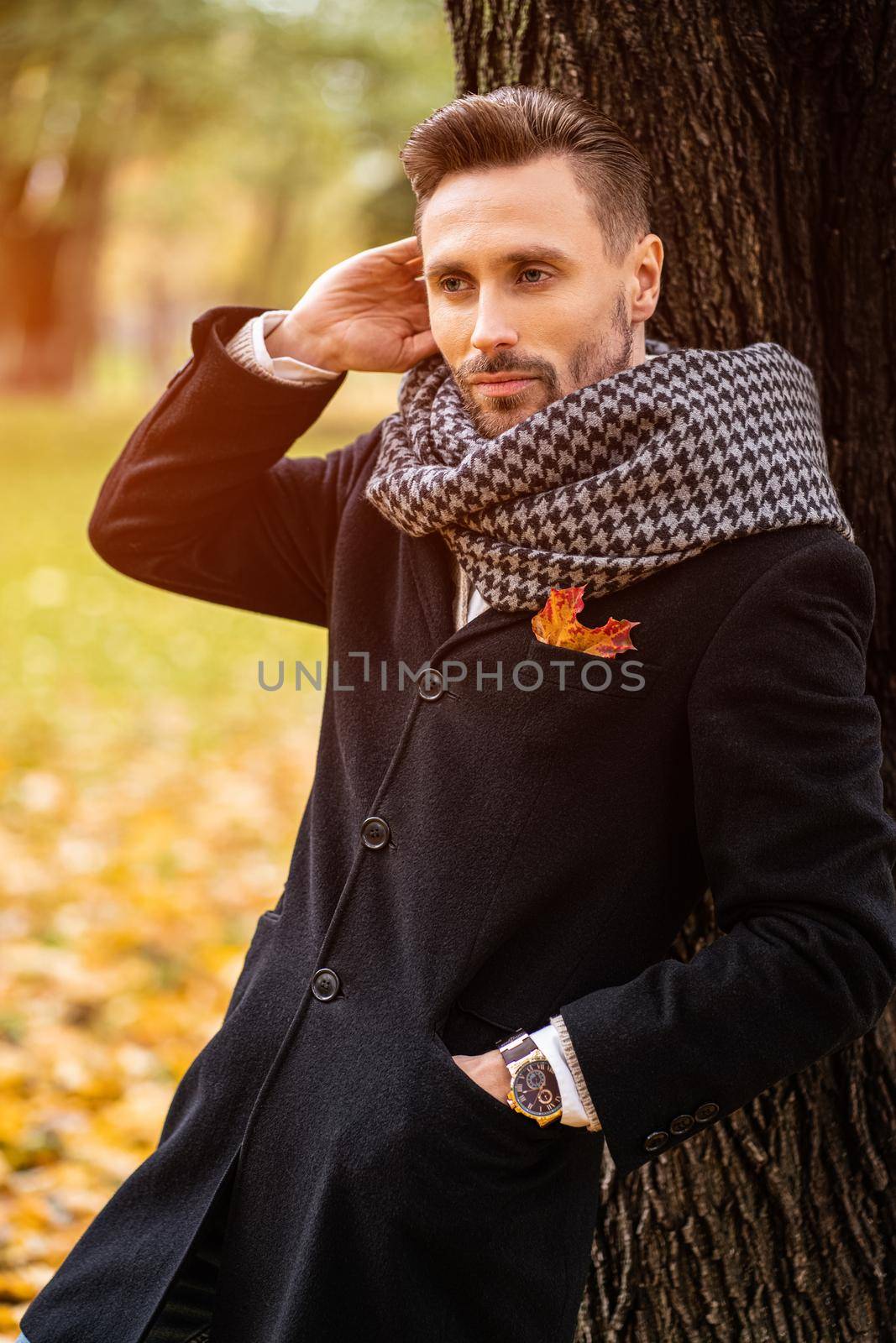 Standing leaned on the tree handsome man straightens his hair wearing dark blue coat. Young freelancer man stands on the street in an autumn coat looking happy waiting foe a date.