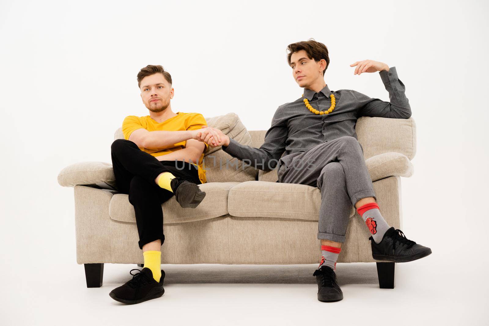 Two cute guys make a bet hand in hand sitting on a white sofa. Group of friends are sitting on a soft couch and communicates isolated on white background.