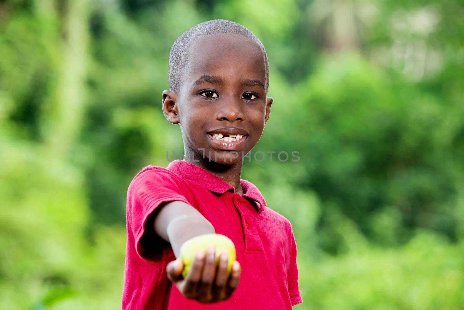 portrait of little cute funny boy standing looking at camera smiling and having hand outstretched with apple. Horizontal color photography.