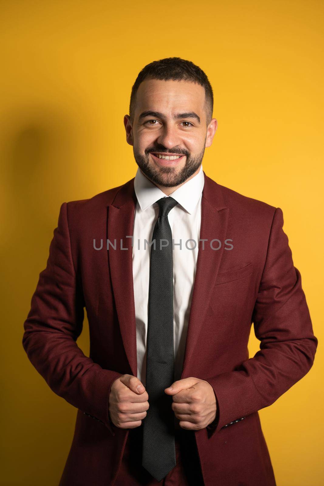 Young handsome man wearing amazing elegant burgundy suit looking on you smiling with hands holding a jacket isolated on yellow background. Men fashion concept.