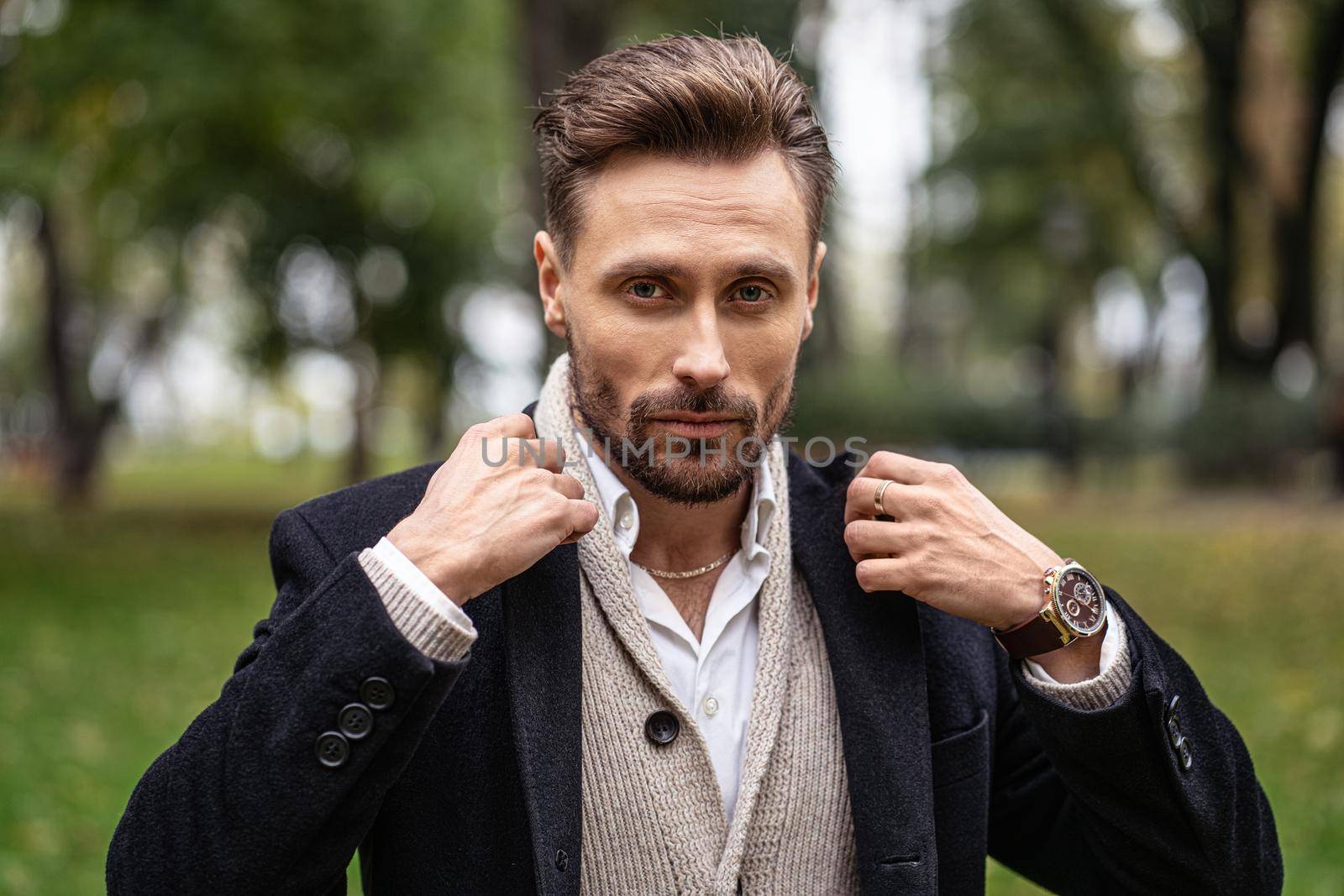 Adjusting the collar of a dark trench coat handsome man standing outdoors in an autumn park smiling looking at the camera. Handsome young man in dark blue coat. Facial expressions, emotions, feelings.