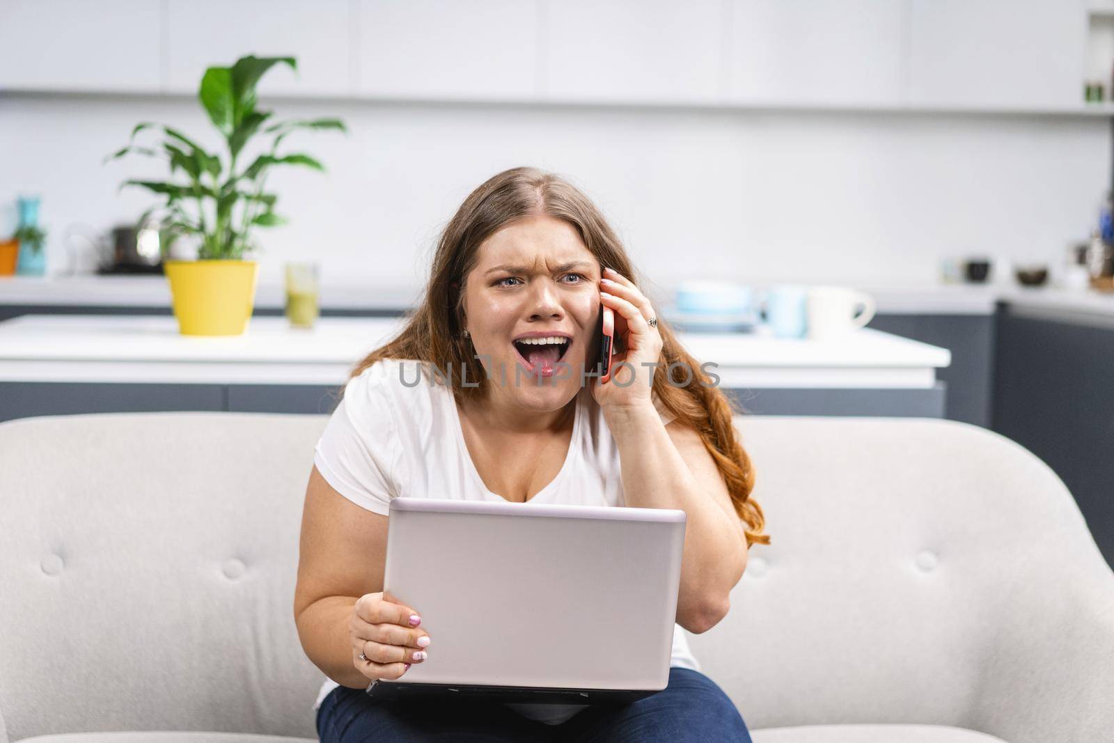 Mad screaming talking on the phone young fat girl working on laptop sitting on the sofa at home with the kitchen on background. Self acceptance. Working distantly from home using laptop by LipikStockMedia