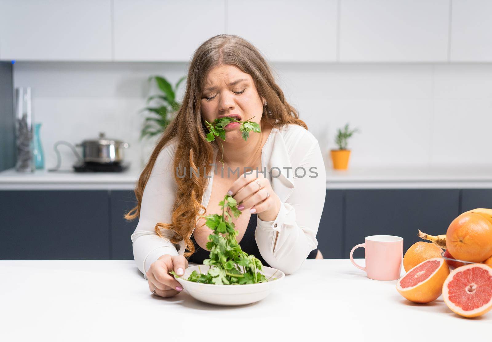 Young girl with weight problems holding fresh salad casting trying to chew it. Curvy body young woman with long blond hair sitting on modern kitchen. Dieting and nutrition concept by LipikStockMedia