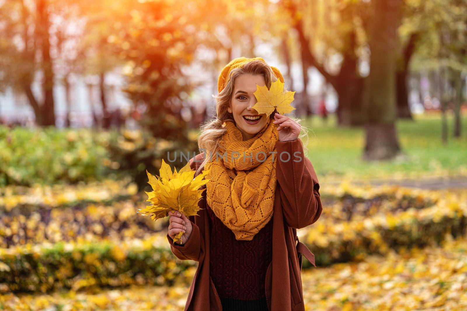 Happy hiding eye woman with a yellowed leaf in yellow knitted beret with autumn leaves in hand and fall yellow garden or park. Beautiful smiling young woman in autumn foliage by LipikStockMedia