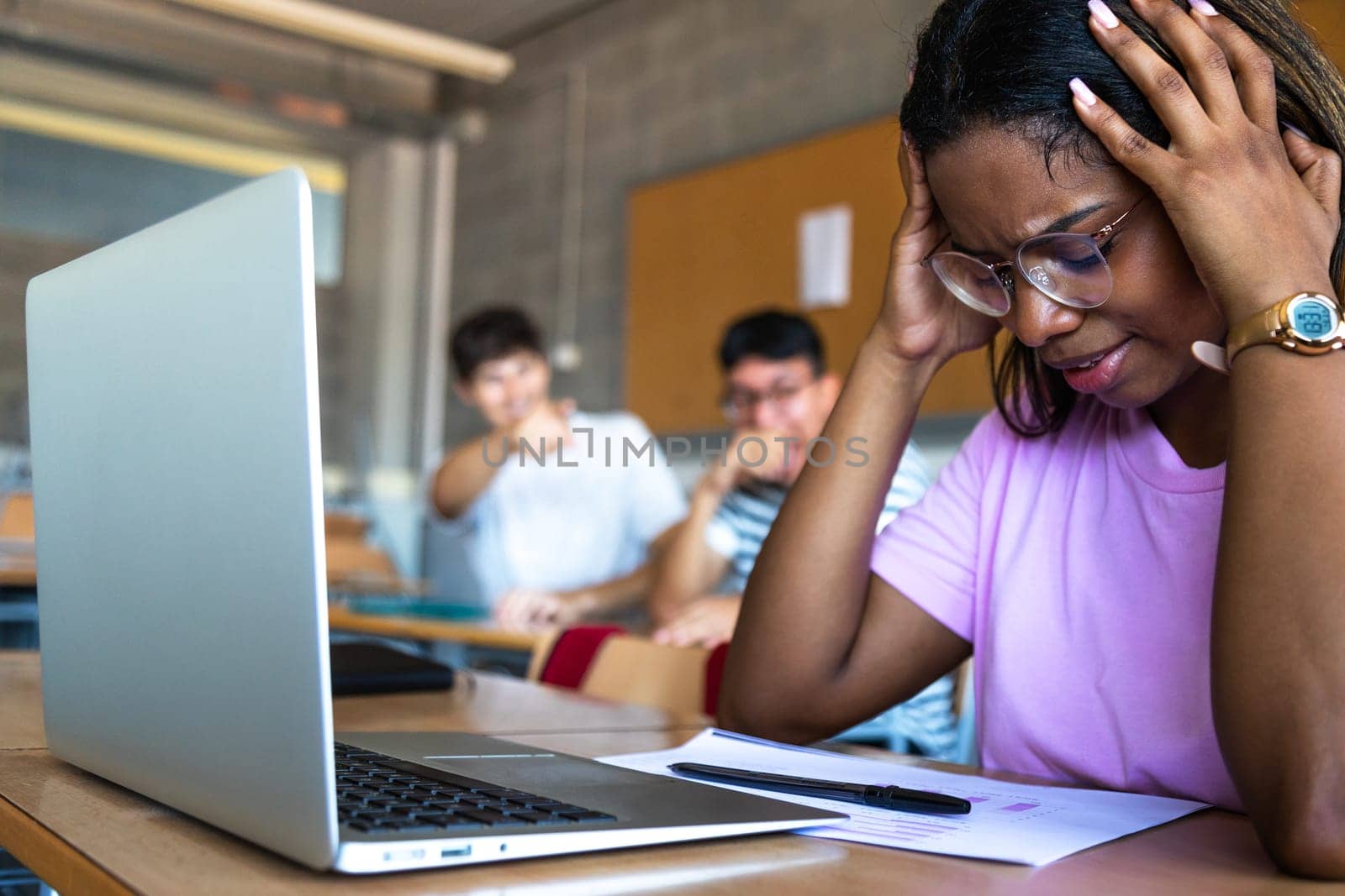 Sad female black teen high school student being bullied by classmates in class. Focus on foreground. Bullying. by Hoverstock