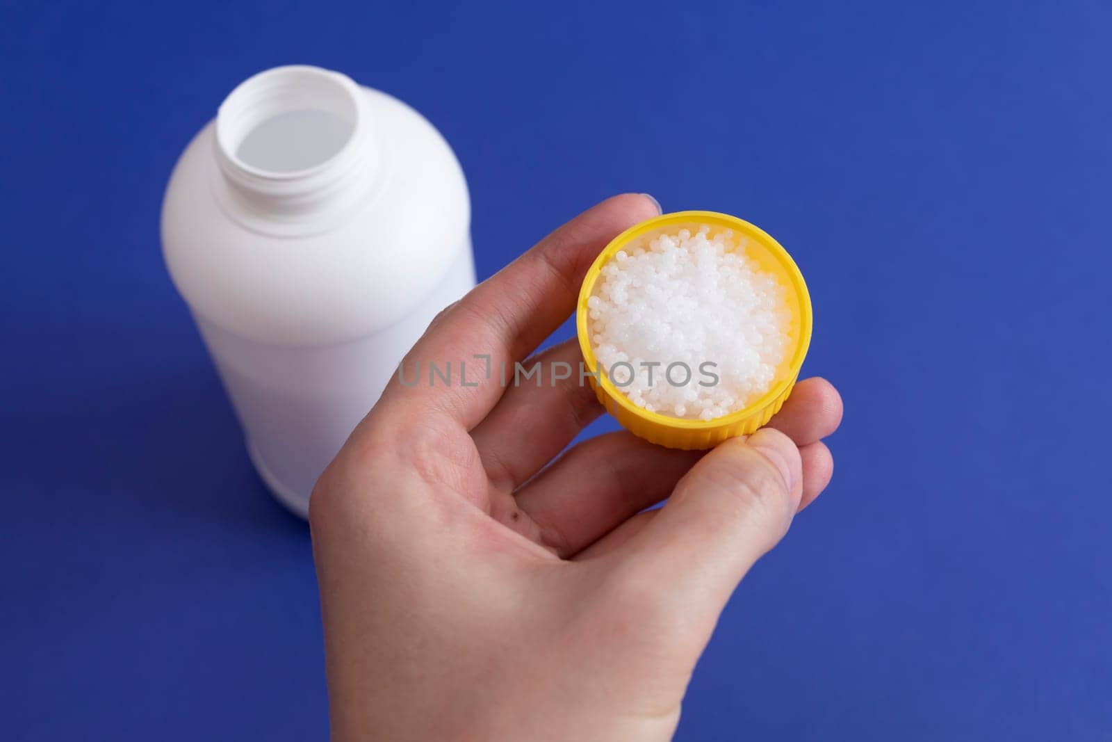 Closeup White Plastic Bottle, Cap Filled With Pipe Cleaner Granules In Hand. Removal Of Blockage In Sink, Shower With Special Chemicals, Drain Cleaner Powder. Unblocking Clog In Kitchen, Bathroom