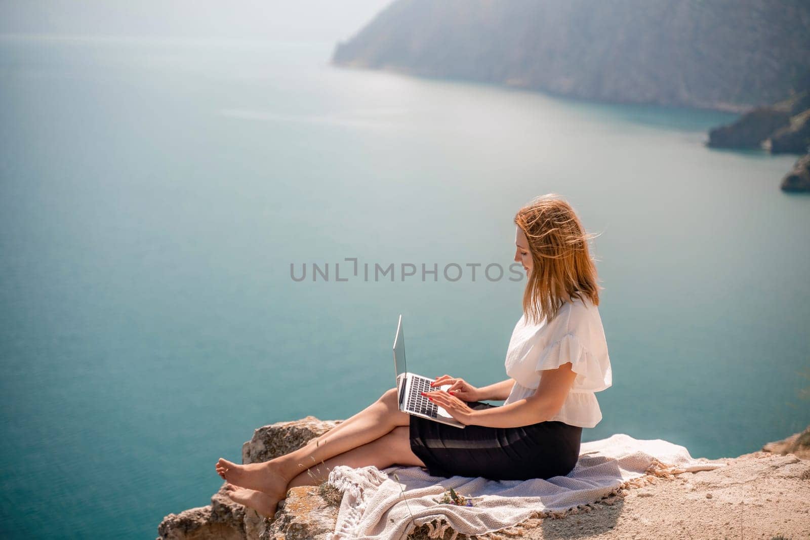 Freelance woman working on a laptop by the sea, typing away on the keyboard while enjoying the beautiful view, highlighting the idea of remote work