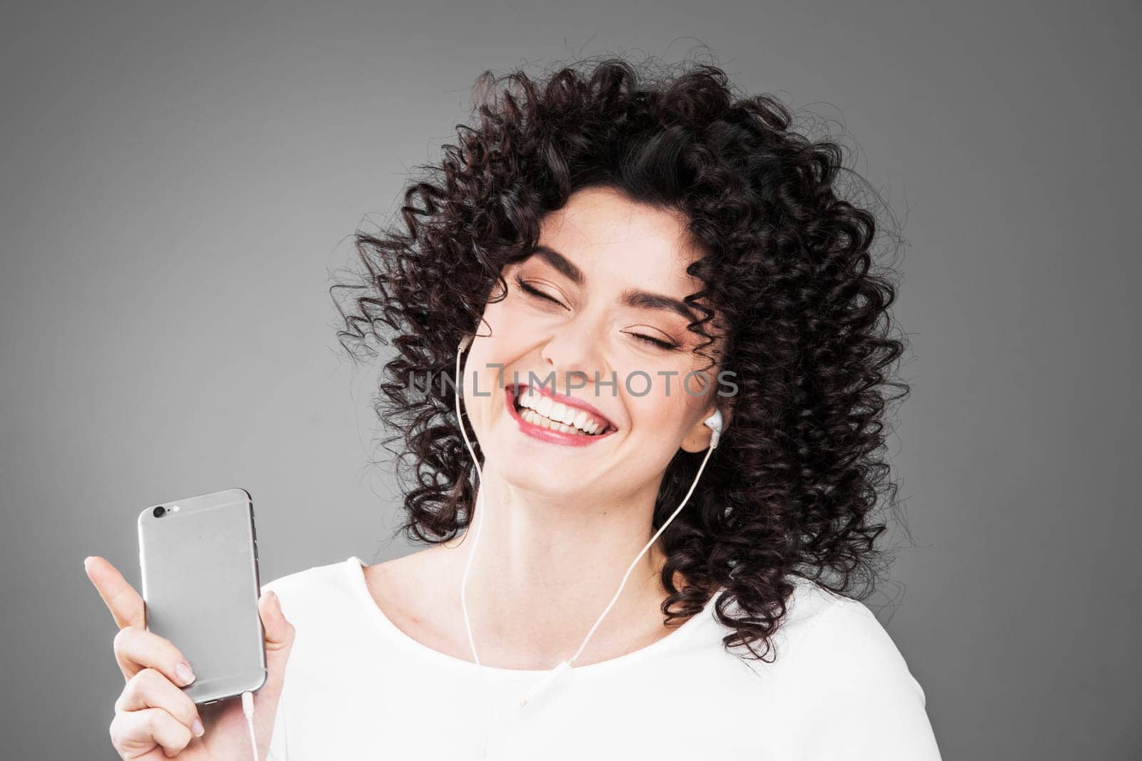 Woman in white dancing with earphones headphones listening to music on phone, gray background